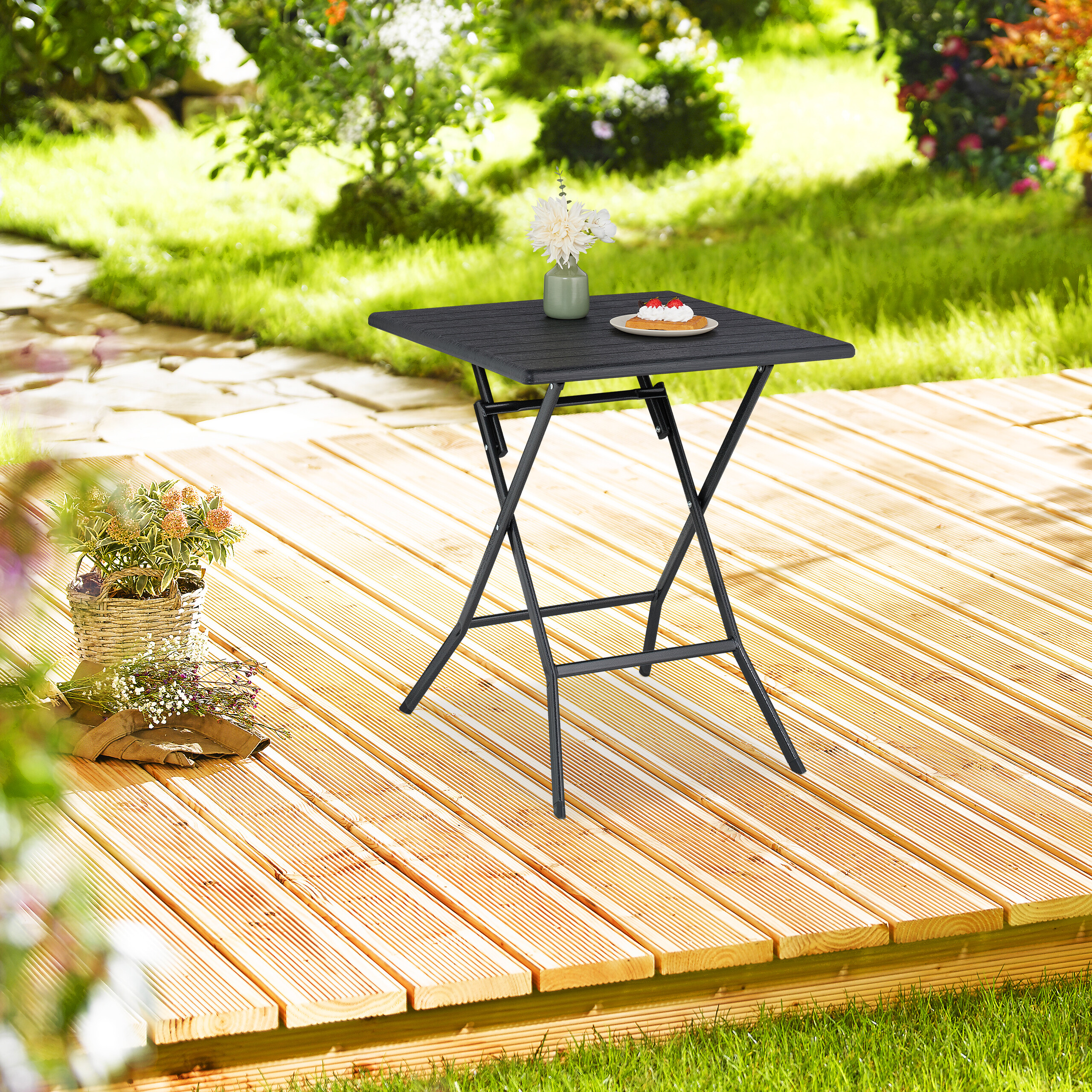 Schwarzer Klapptisch auf einer Holzterrasse im Garten, dekoriert mit einer Blumenvase und Kuchen.