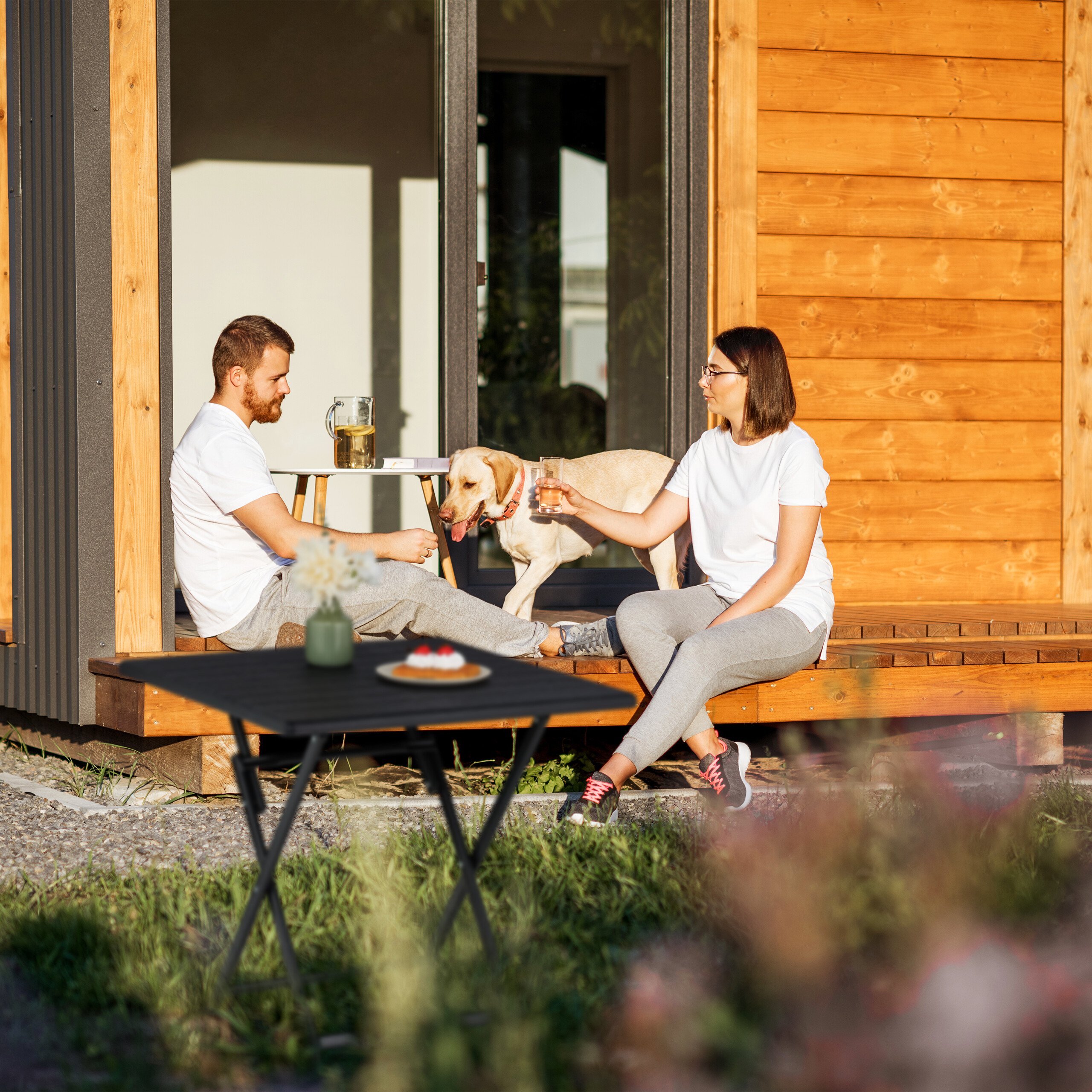 Ein Mann und eine Frau entspannen mit ihrem Golden Retriever auf einer Holzveranda vor einem modernen Haus.