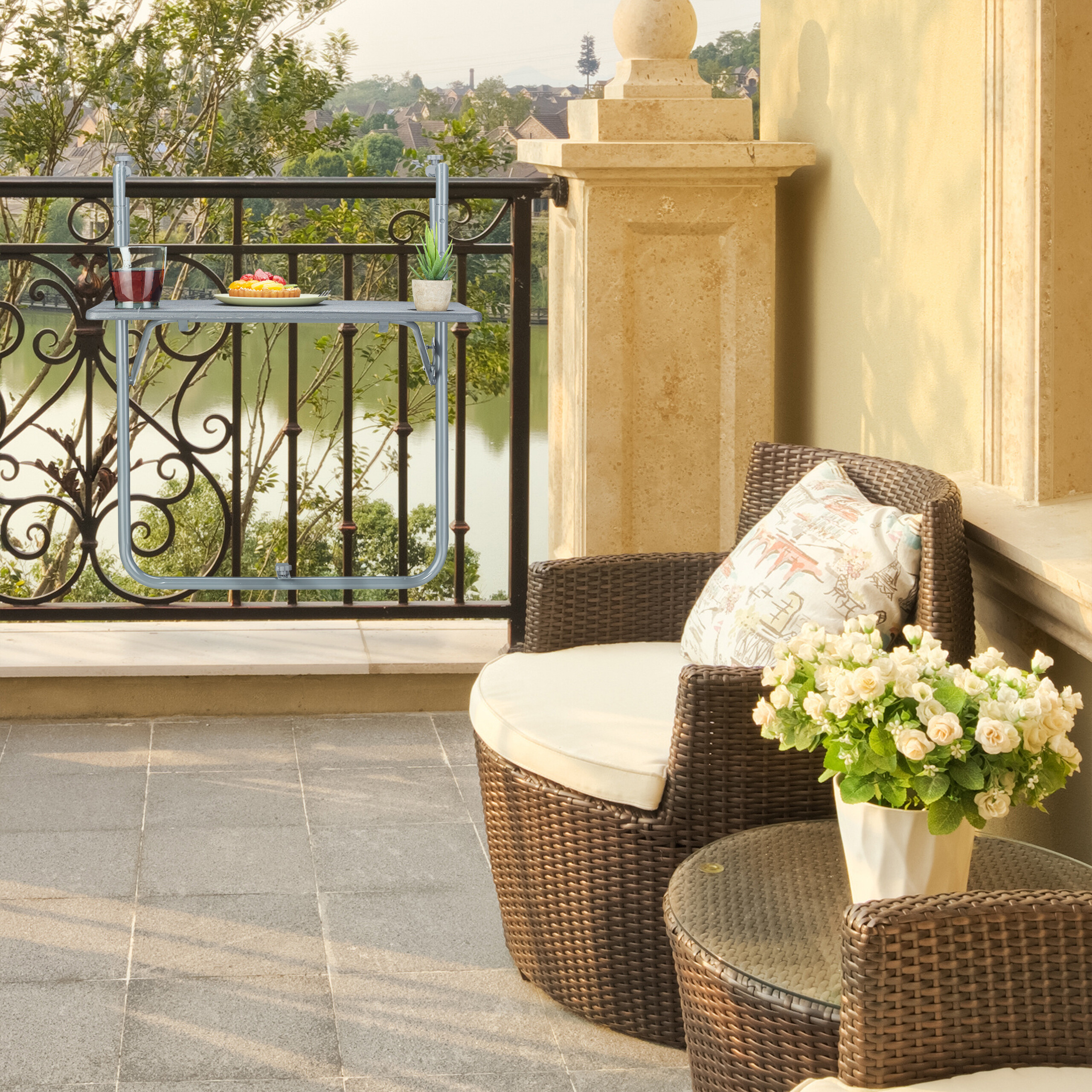 Balkon mit hängendem Tisch am Geländer, Rattansessel, Beistelltisch mit Blumen und Seeblick im Hintergrund.