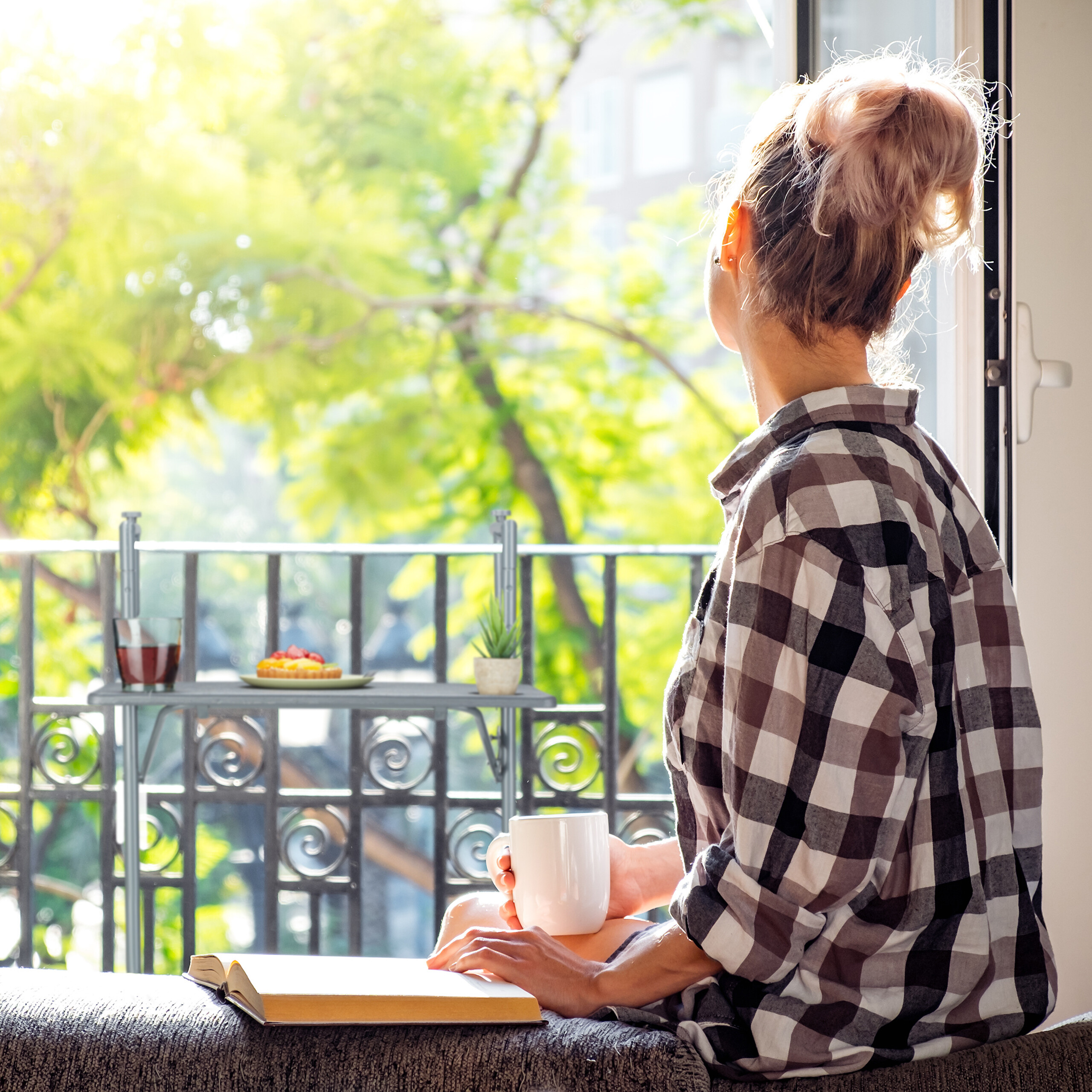 Eine Frau in kariertem Hemd sitzt am Fenster mit Kaffeetasse und Buch, im Hintergrund sonnige Bäume.