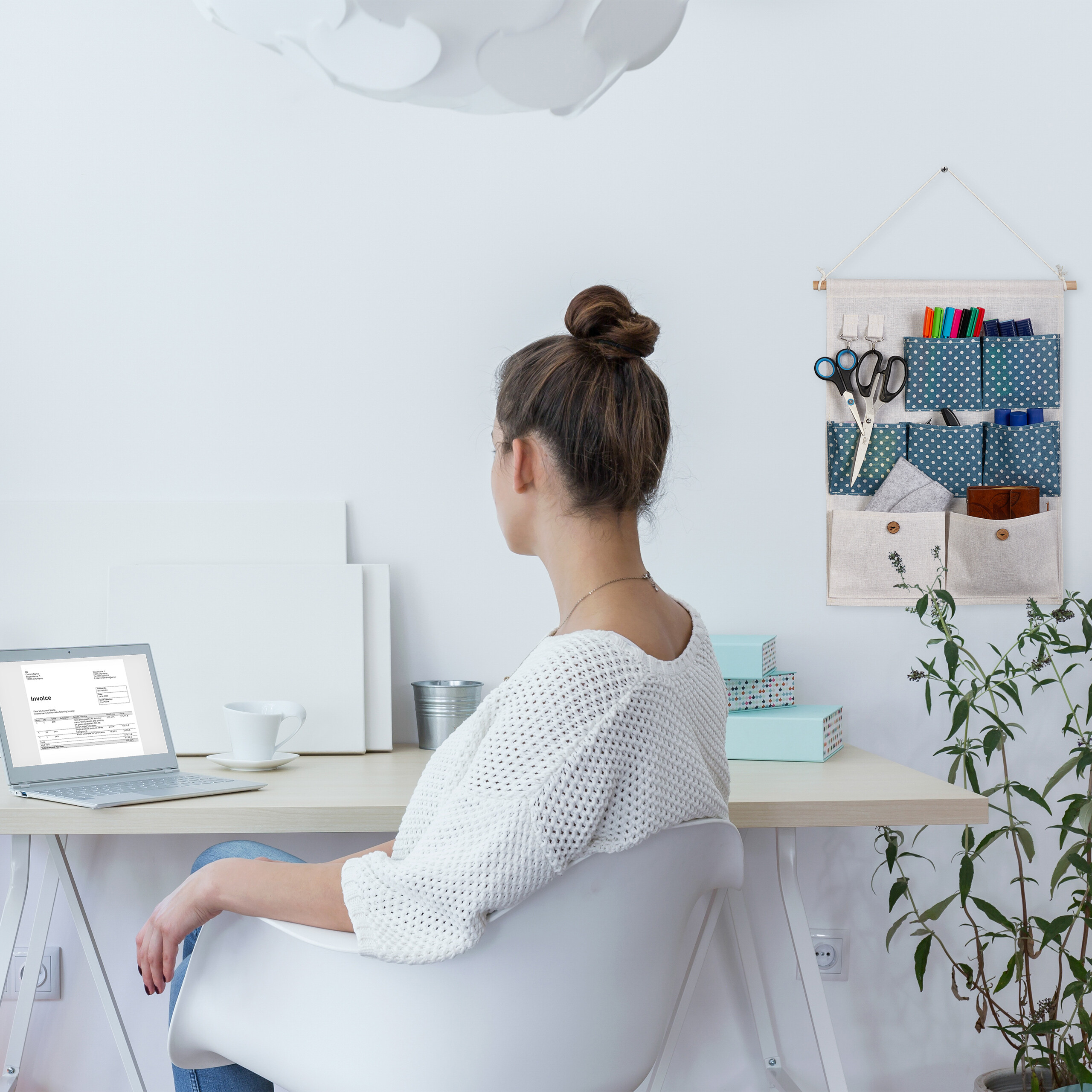 Eine Frau sitzt am Schreibtisch vor einem Laptop, während ein Wandorganizer und eine Pflanze im modernen Raum zu sehen sind.