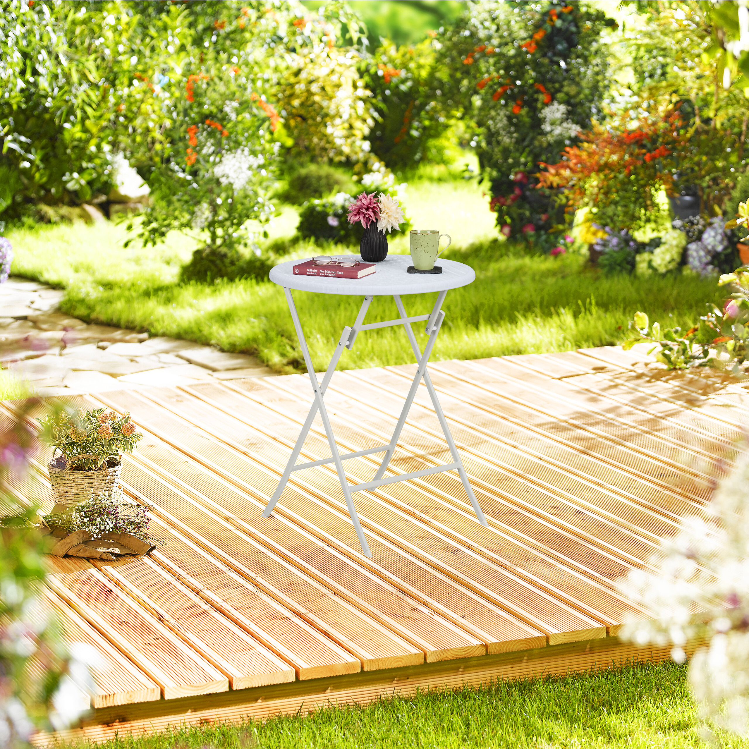 Ein kleiner weißer Klapptisch auf einer Holzterrasse in einem sonnigen Garten mit Blumen und Tasse.