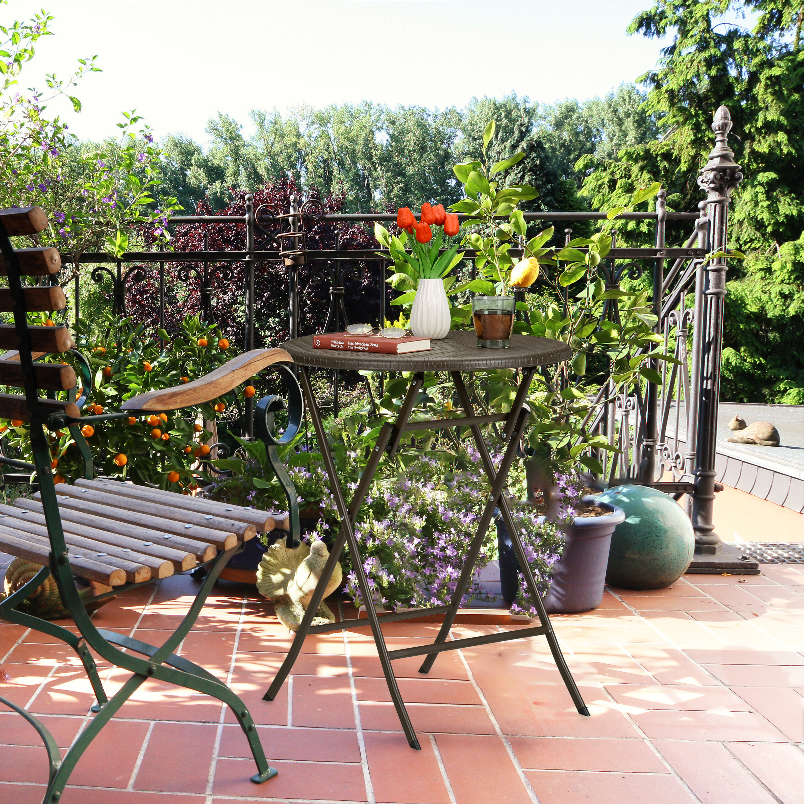 Sonnige Terrasse mit Tisch, Tulpen in Vase, Buch, Getränk, Stuhl und vielen grünen Pflanzen auf Terrakottaböden.