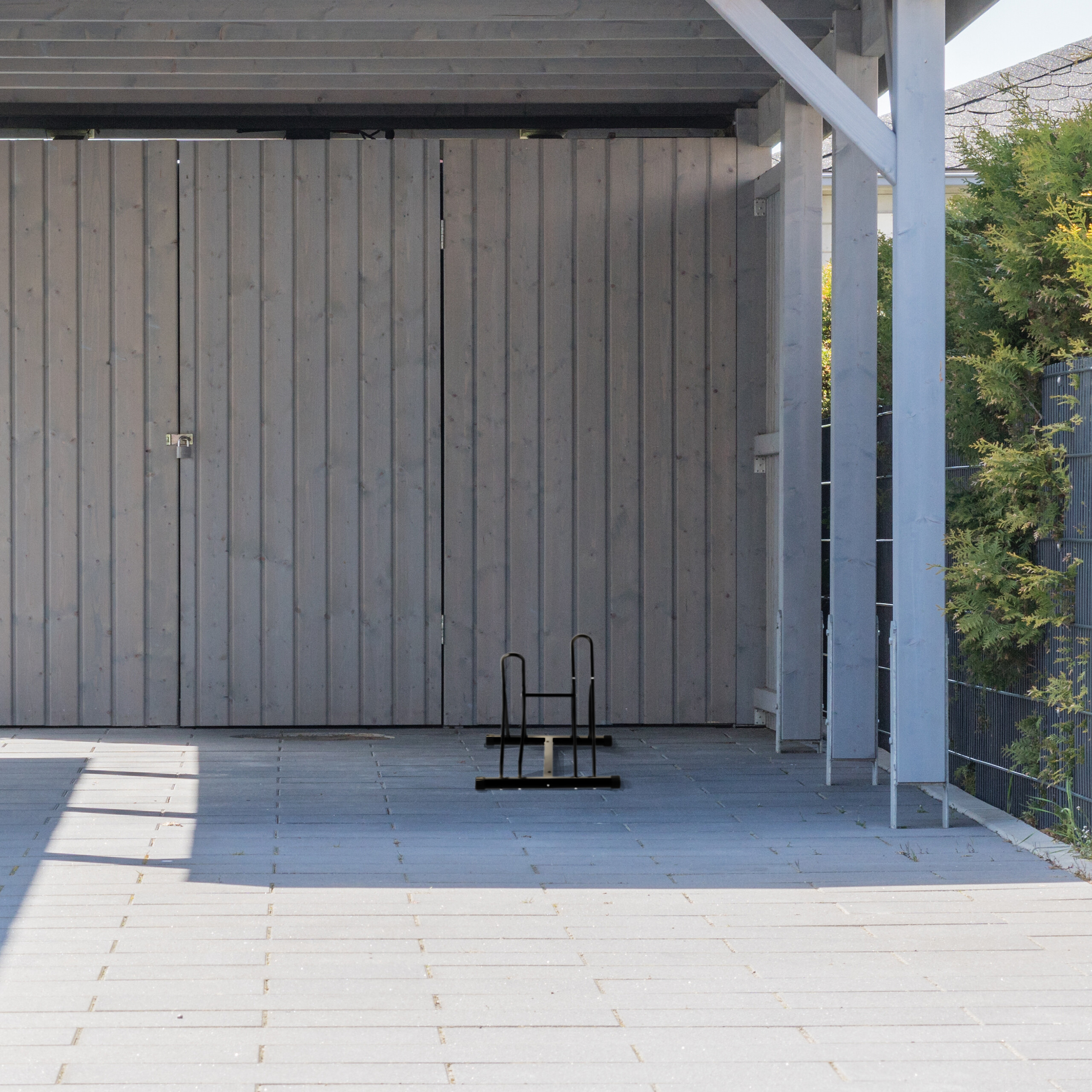 Grauer Carport mit Holztafelwand, gefliester Boden, schwarzer Fahrradständer und grüne Büsche im Hintergrund.