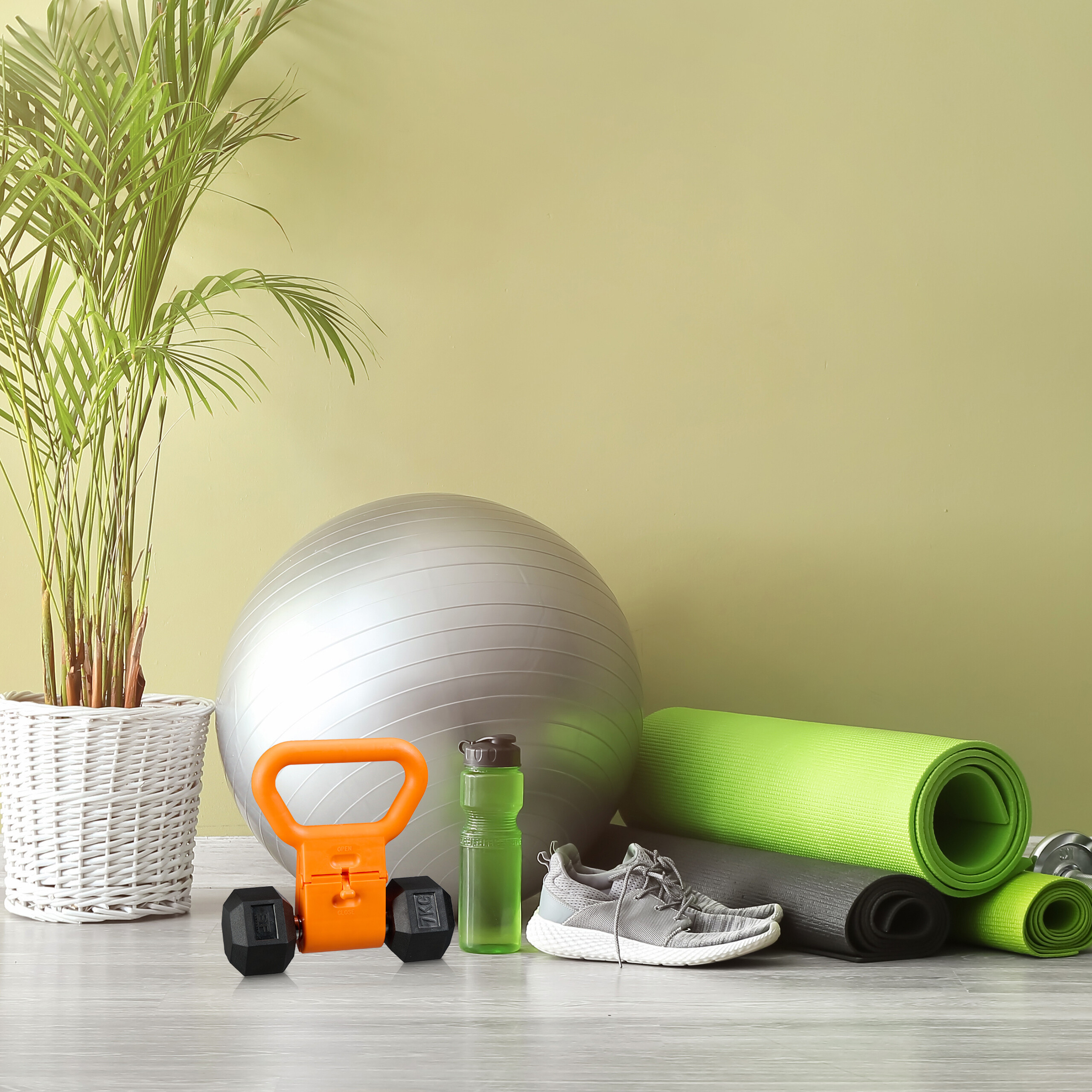 Fitnessgeräte wie Gymnastikball, Kettlebell, Yogamatten und Turnschuhe vor einer hellen Wand.