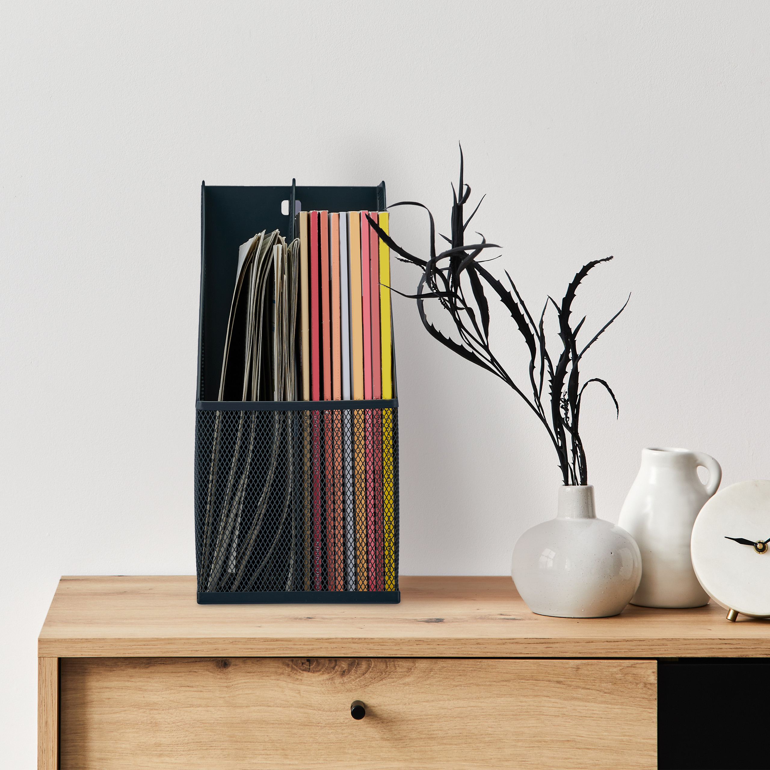 Ein schwarzer Metallorganizer mit bunten Büchern steht auf einem Holzschrank, neben zwei weißen Vasen und einer Uhr.