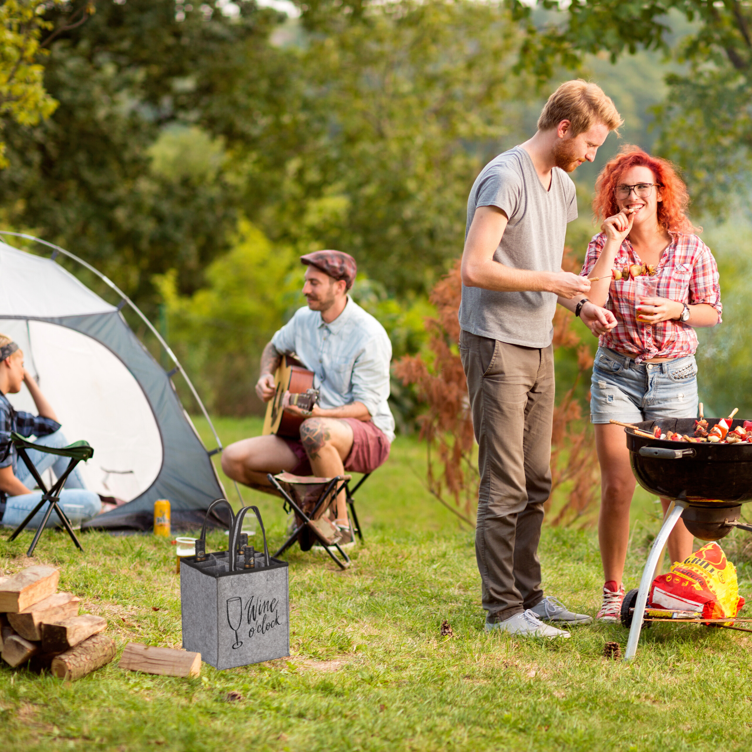 Freunde beim Grillen und Gitarrespielen auf einem Campingplatz, mit einem Zelt und einer Weintasche im Vordergrund.