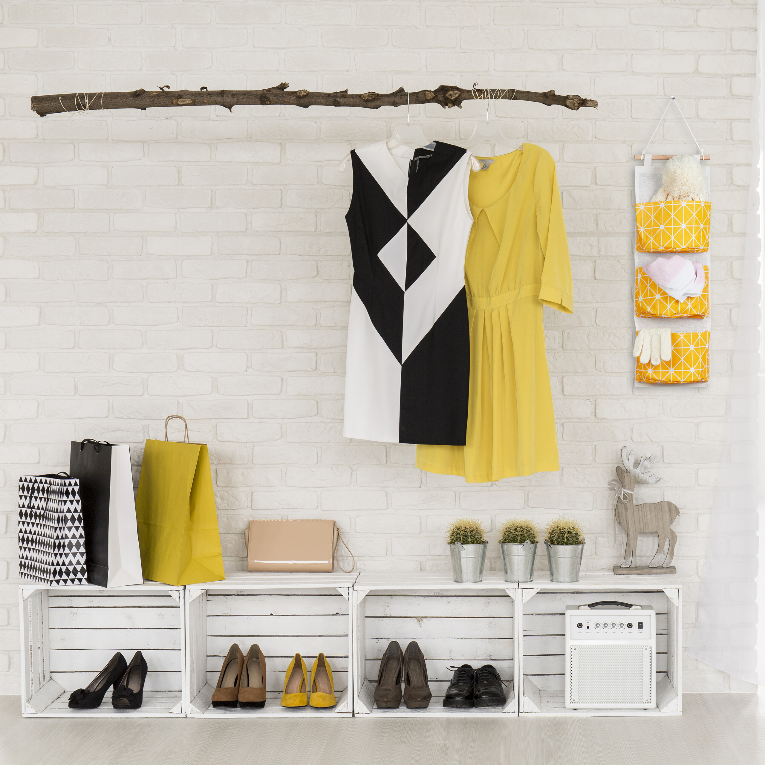 Kleidung hängt an einem Ast vor einer Backsteinwand, darunter Holzkisten mit Schuhen und Taschen.
