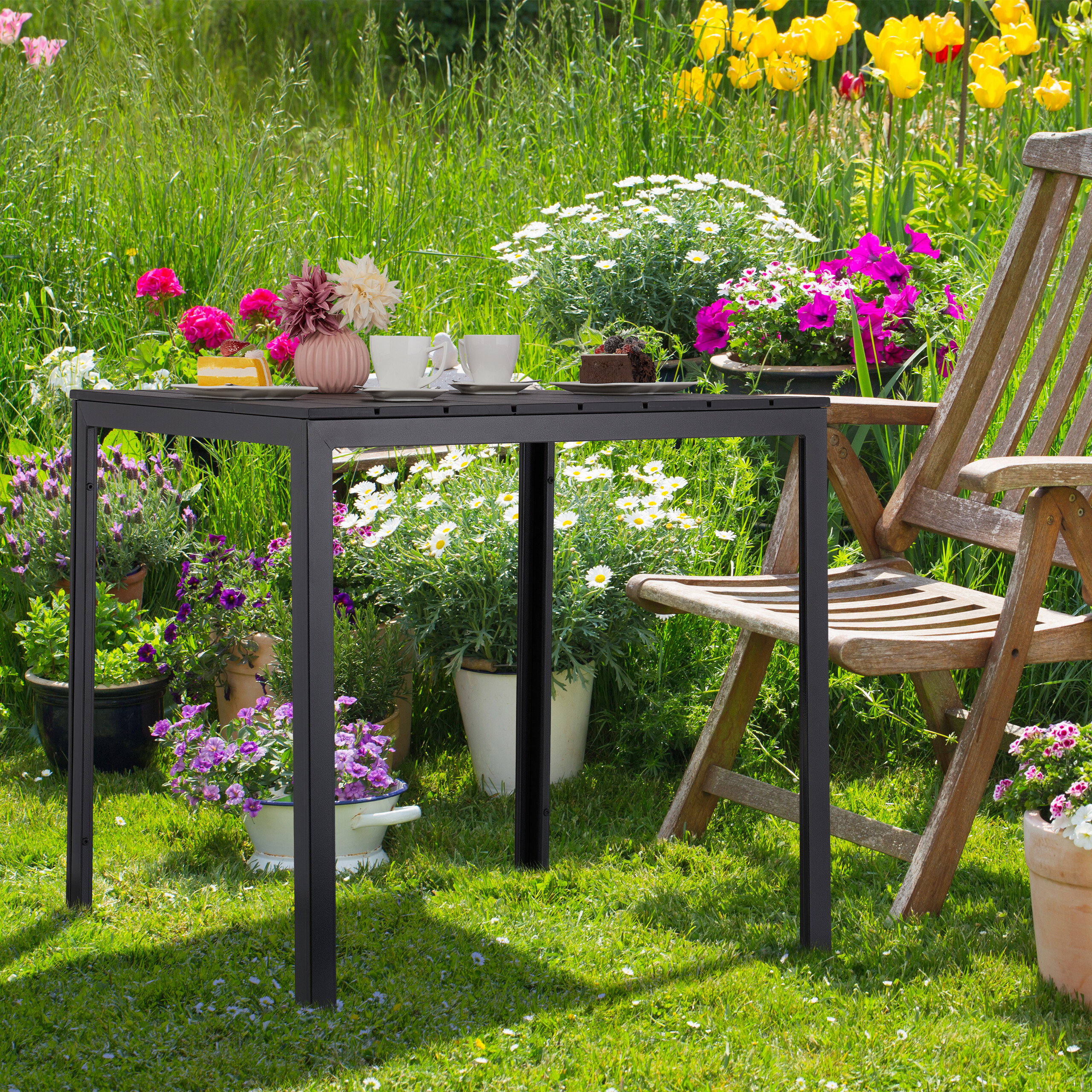 Ein schwarzer Tisch mit Kaffeetassen und Kuchen steht auf einer grünen Wiese im Garten mit Blumen.