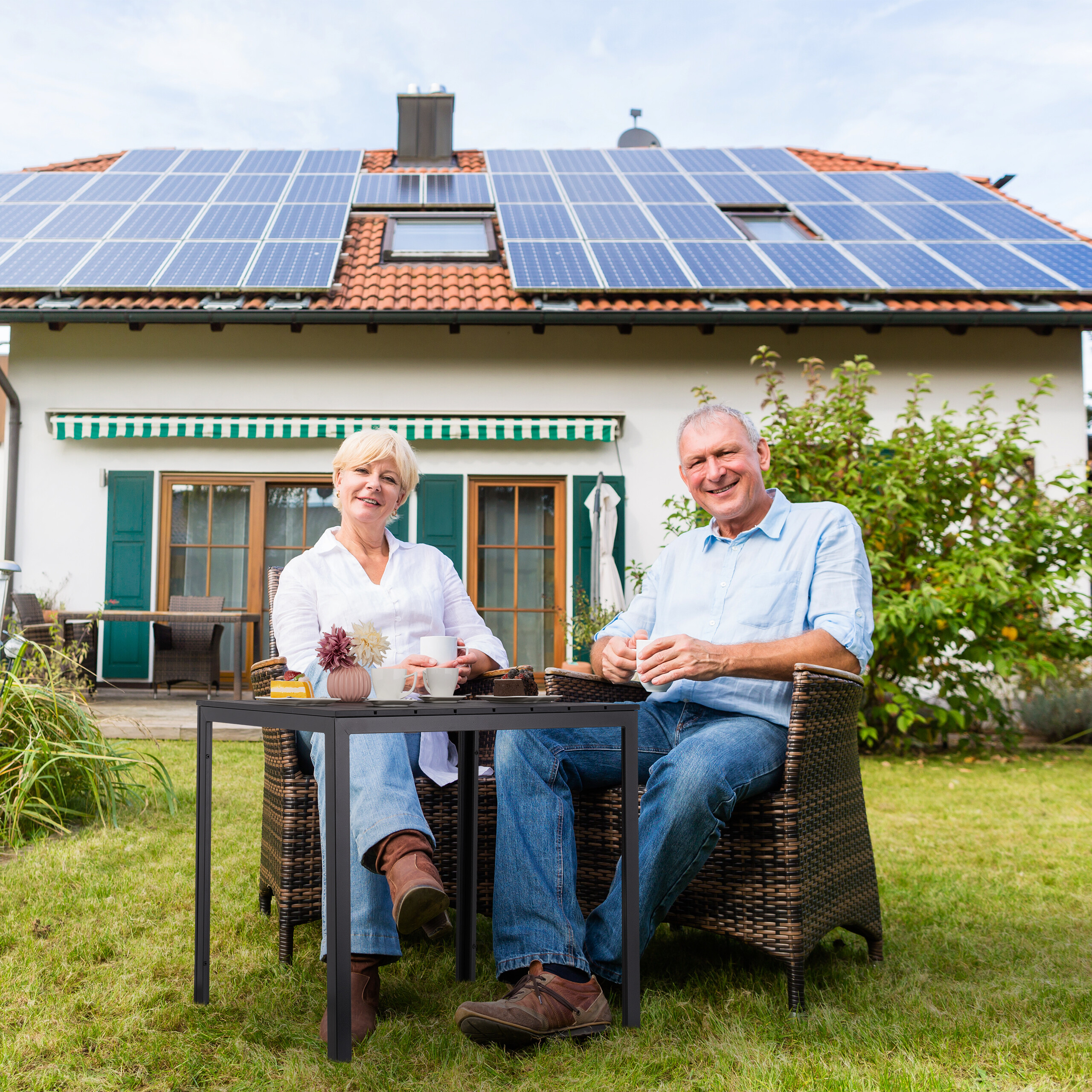 Älteres Paar sitzt im Garten vor einem Haus mit Solarzellen auf dem Dach.