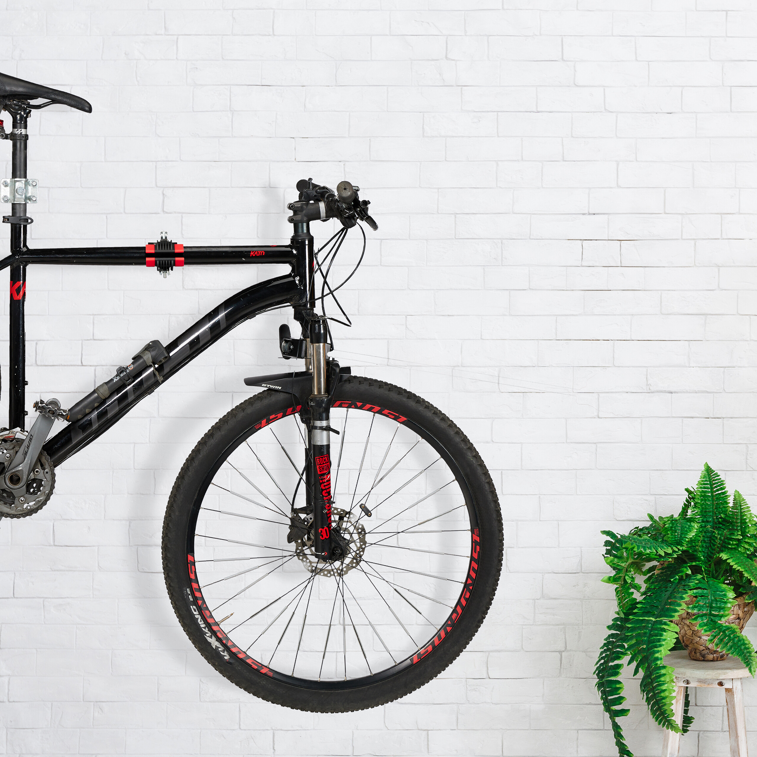 Ein schwarzes Mountainbike hängt an einer weißen Backsteinwand, rechts daneben steht eine grüne Topfpflanze auf einem Hocker.