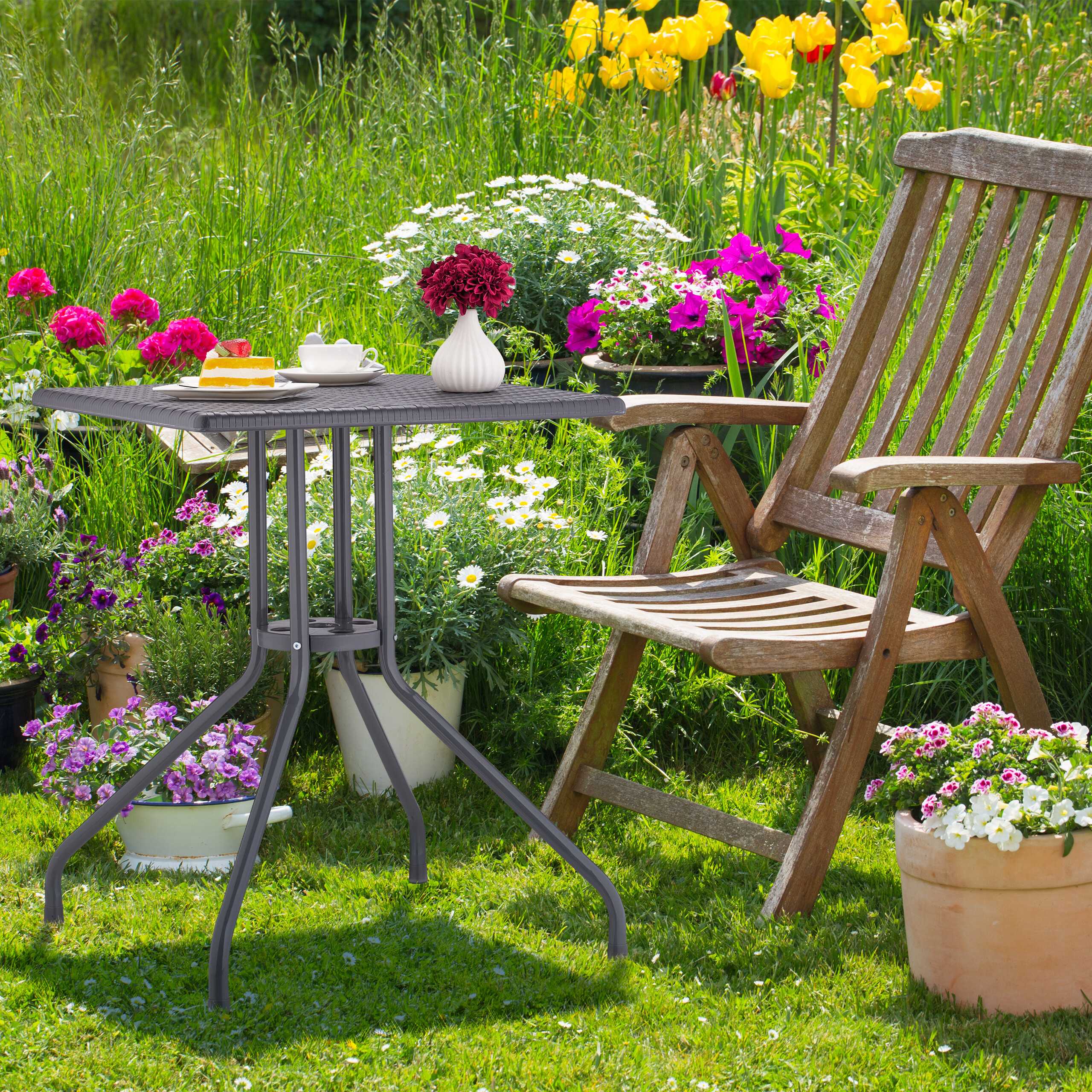 Ein Holzstuhl und ein kleiner dunkler Tisch mit Kuchen und Blumenvase im üppigen Garten.