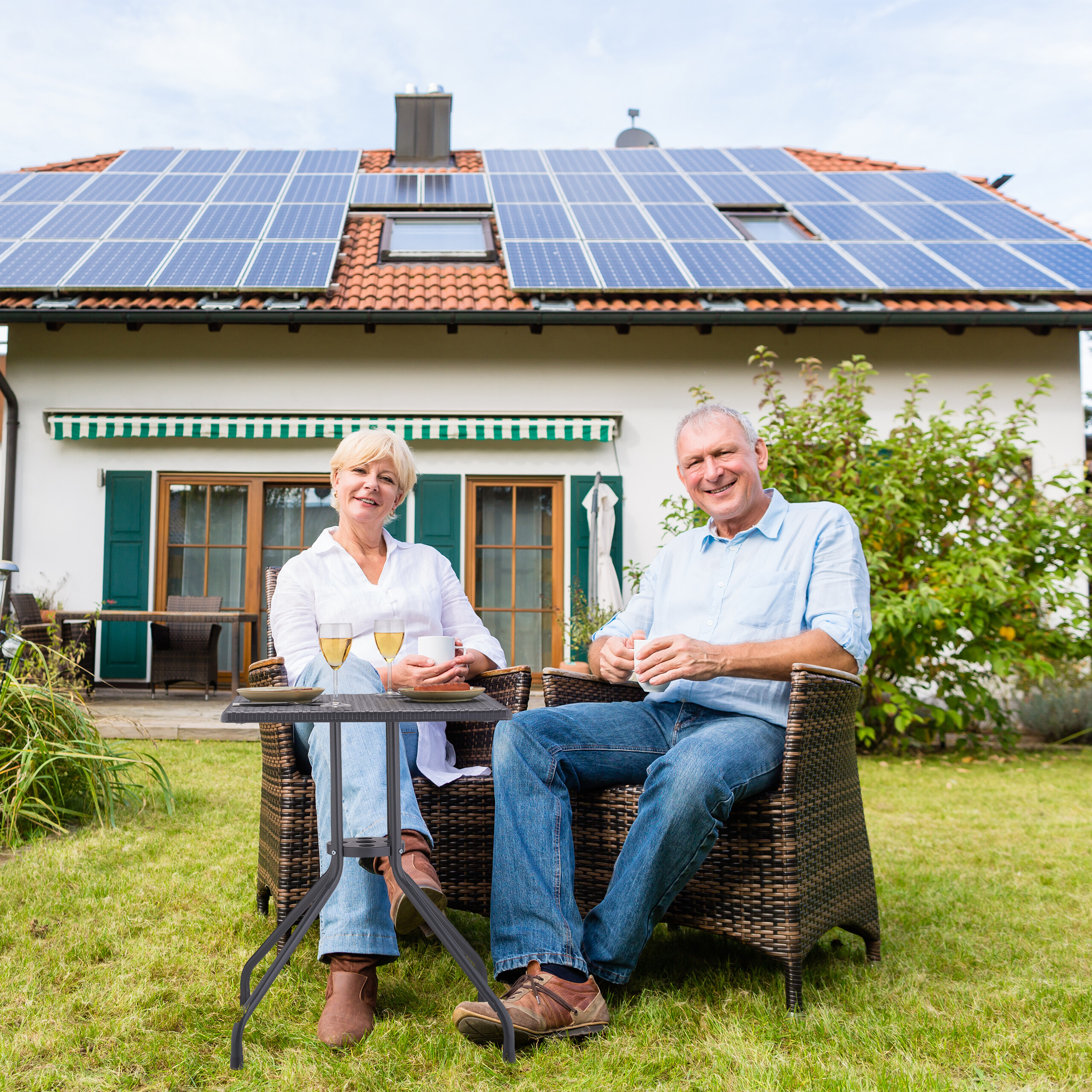 Älteres Paar sitzt entspannt im Garten vor einem Haus mit Solaranlage.