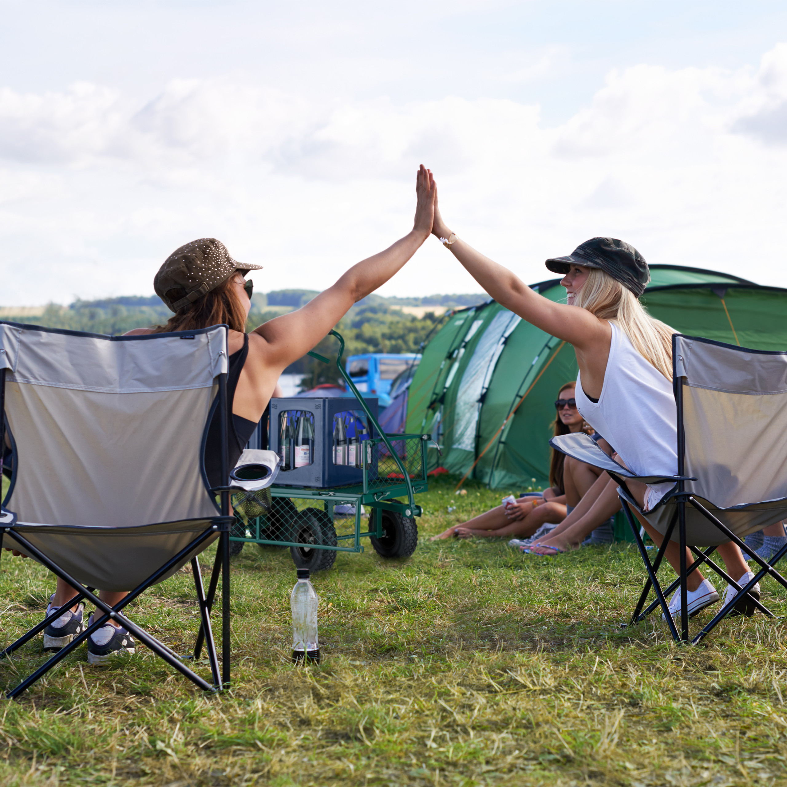 Zwei Frauen sitzen in Campingstühlen und geben sich High Five vor grünen Zelten.