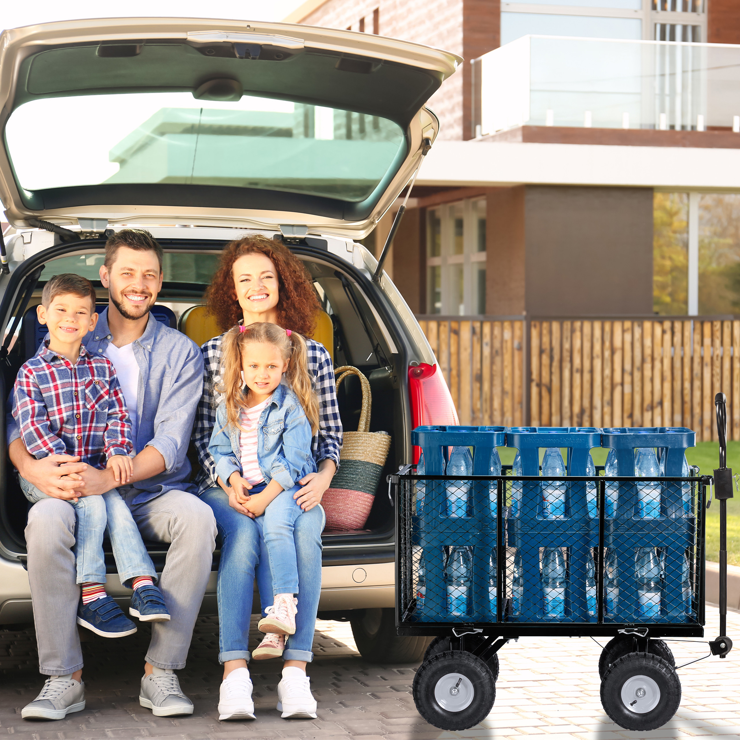 Eine lächelnde vierköpfige Familie sitzt im offenen Kofferraum neben einem Bollerwagen mit Wasserflaschen.