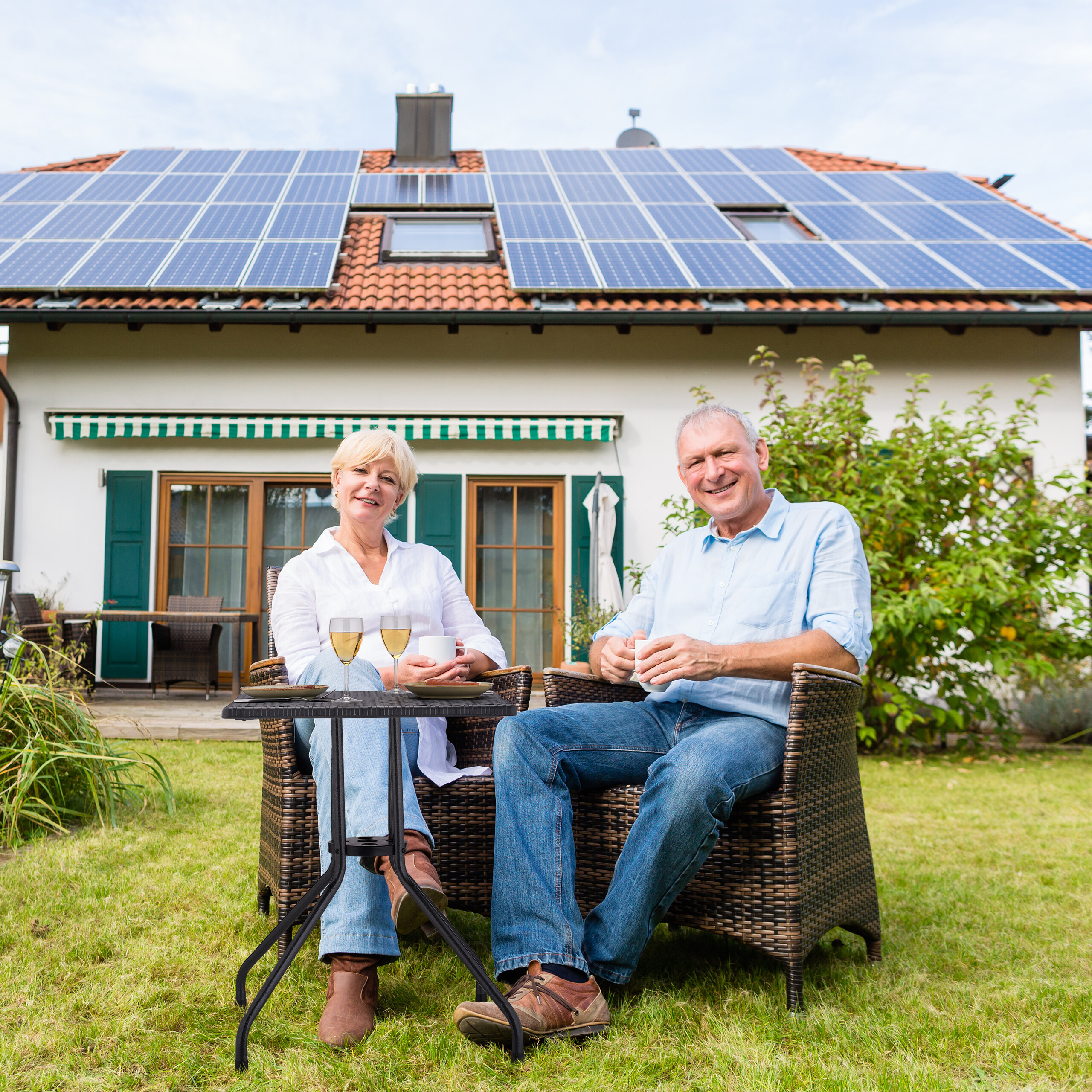 Älteres Paar sitzt im Garten vor einem Haus mit Solaranlage.