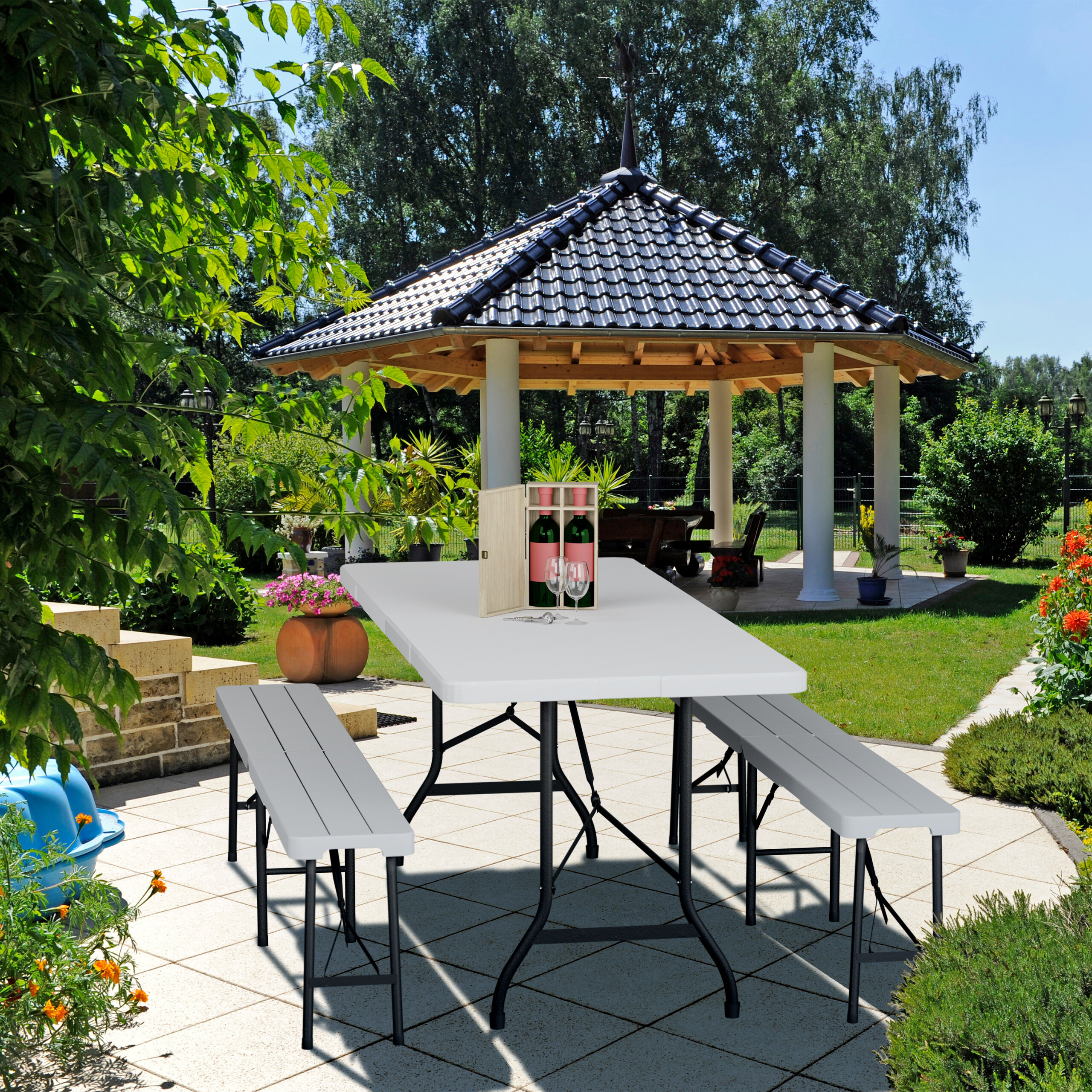 Ein weißer Tisch mit Bänken auf einer Terrasse, dahinter ein Pavillon und ein Garten.