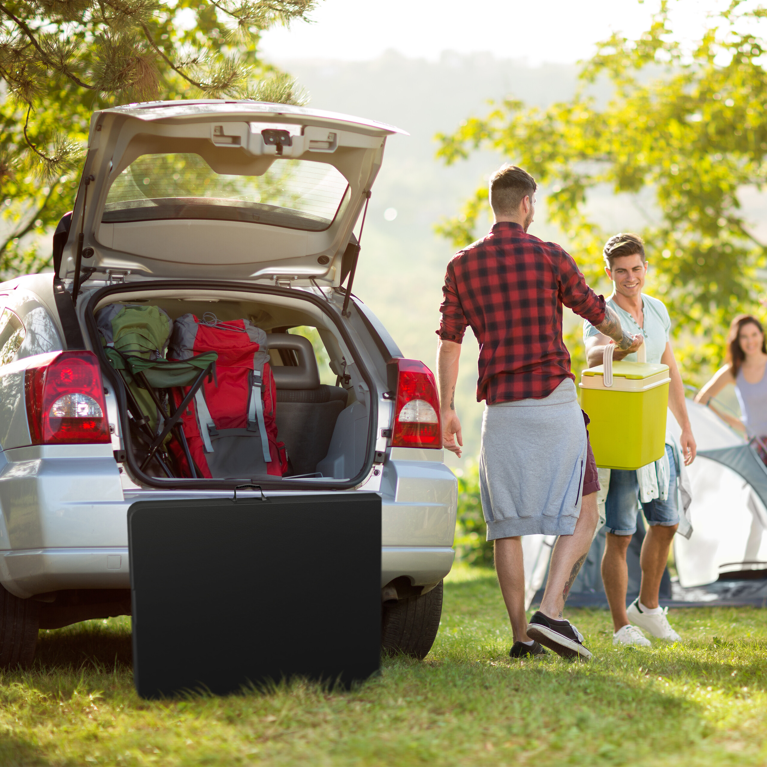 Freunde beim Camping laden Ausrüstung aus dem Kofferraum eines Autos, daneben steht ein Zelt.