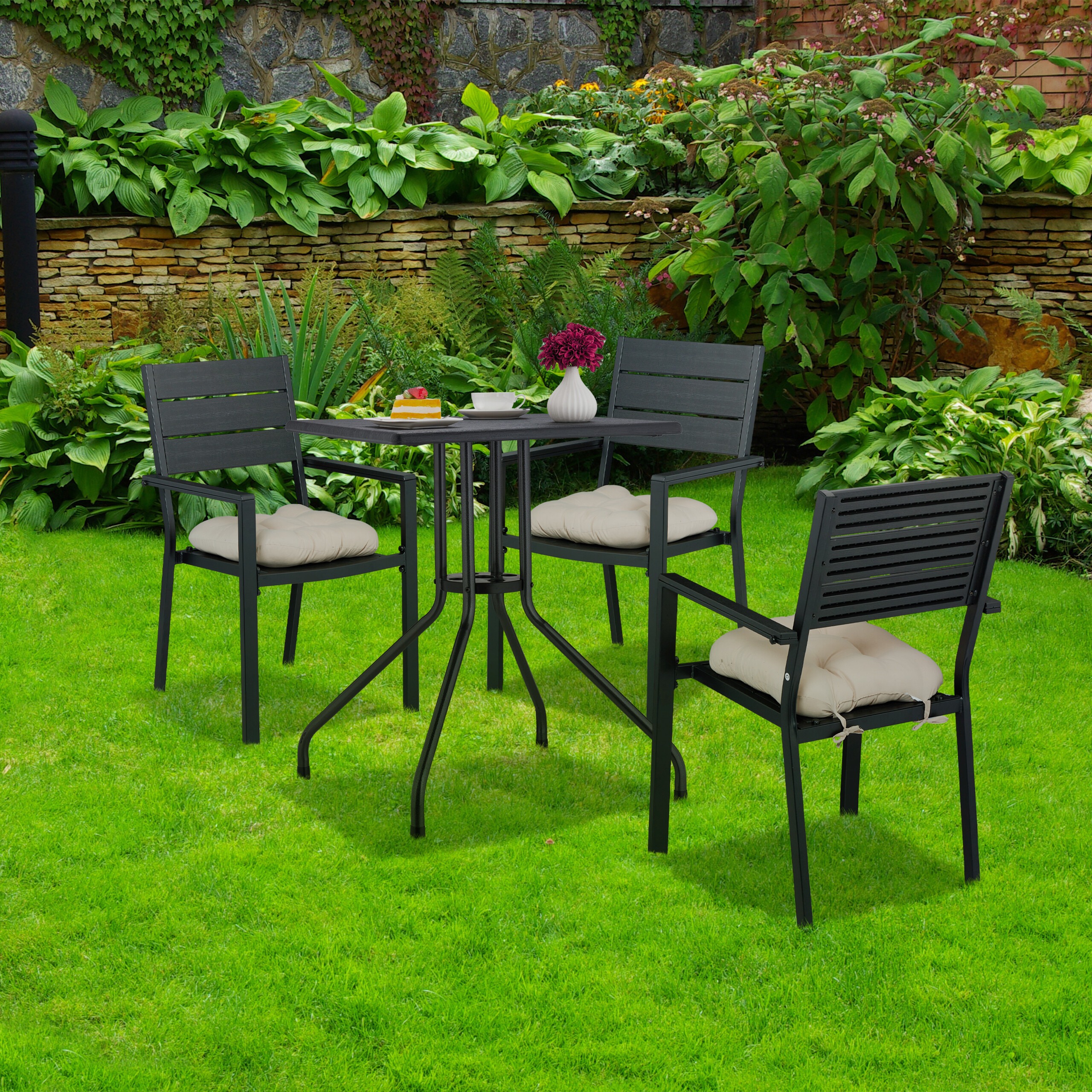 Ein schwarzer Gartentisch mit drei Stühlen und Kissen auf grünem Rasen vor einer Steinwand.