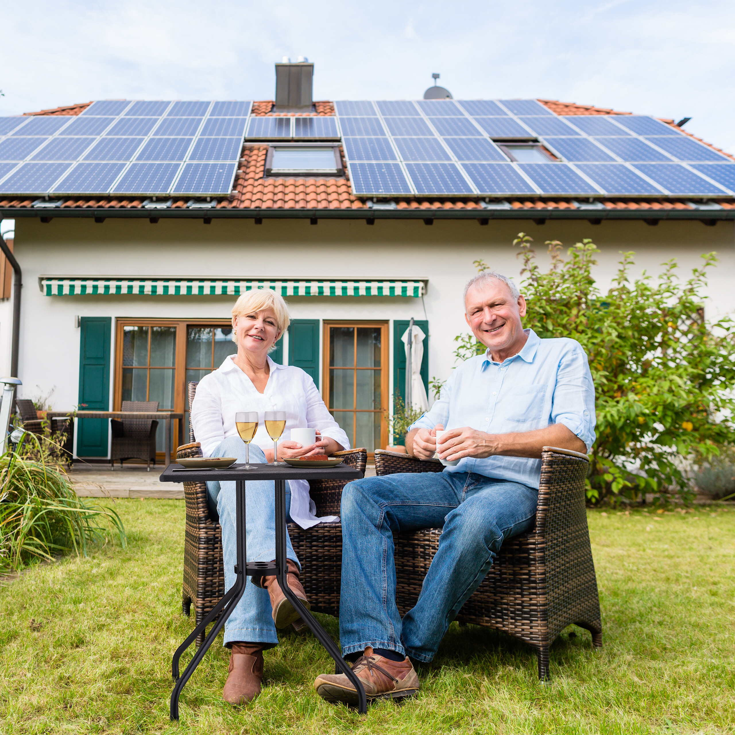 Älteres Paar sitzt im Garten vor einem Haus mit Solaranlage.