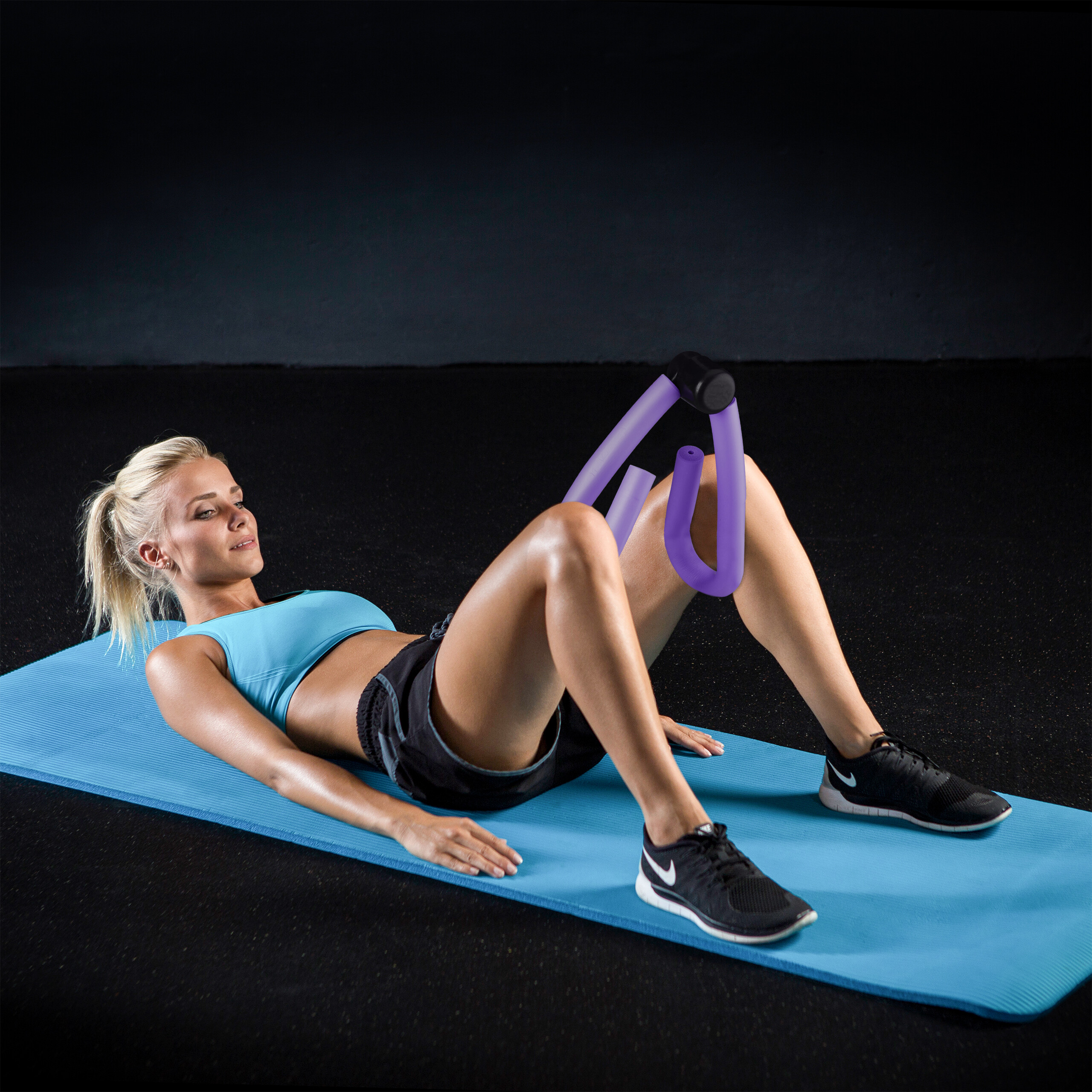 Eine blonde Frau in blauem Sport-BH und schwarzen Shorts benutzt einen violetten Oberschenkel-Trainer auf einer blauen Yogamatte.