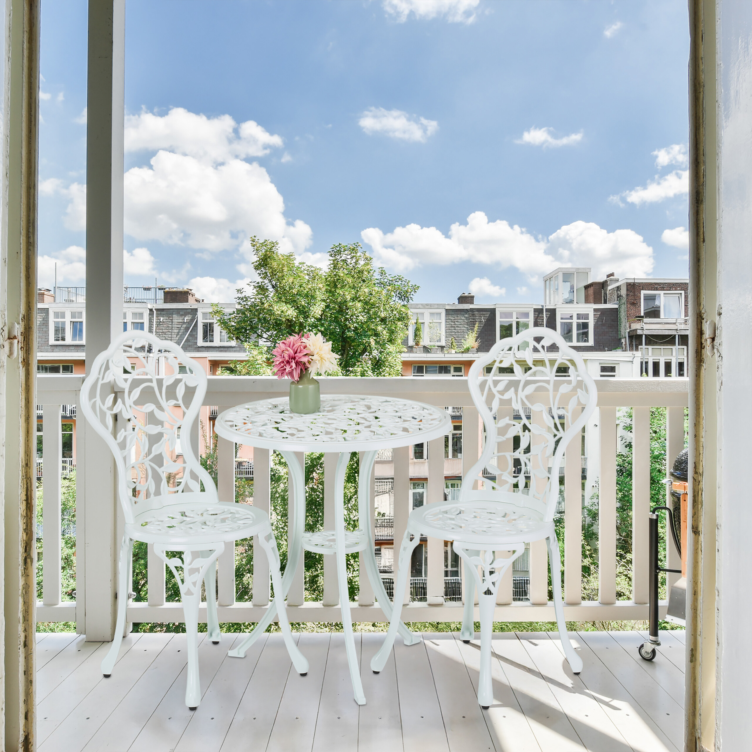 Ein weißer Metalltisch mit zwei Stühlen und Blumen steht auf einem sonnigen Balkon mit Blick auf Gebäude.