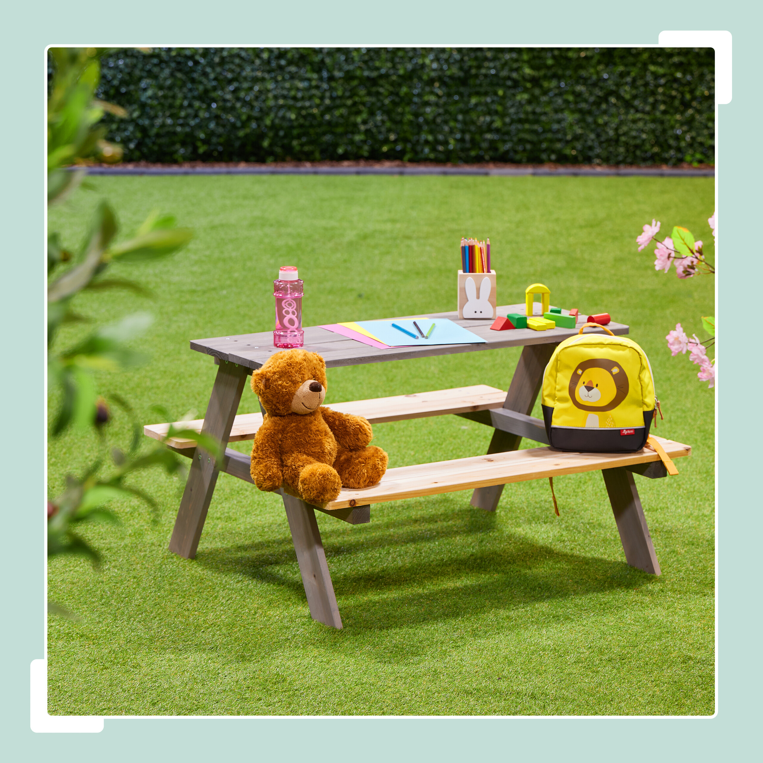 Ein hölzerner Picknicktisch auf Rasen mit Teddybär, Rucksack, Trinkflasche, Stiften und Papier.