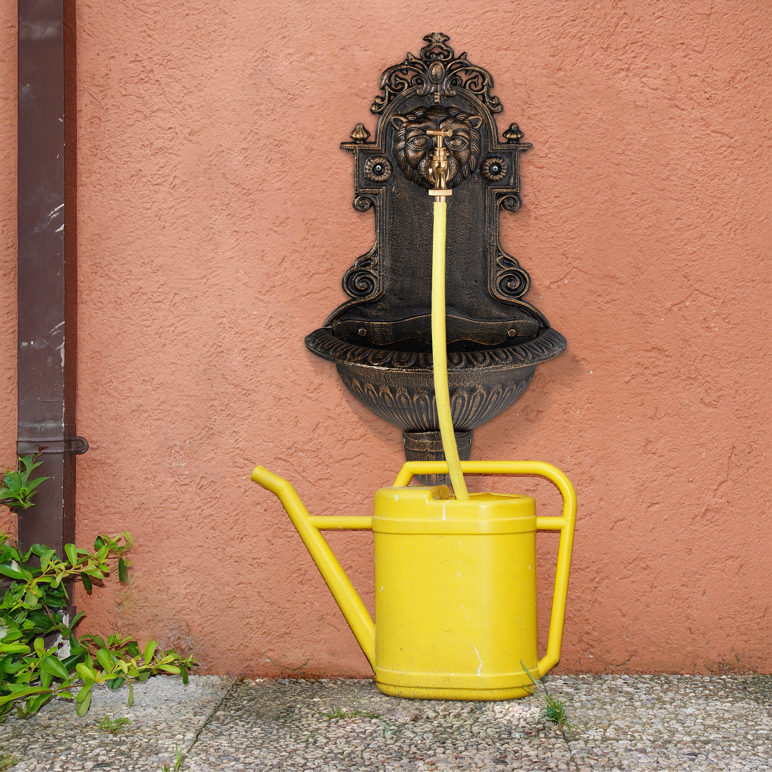 Ein antiker bronzefarbener Wandbrunnen an einer rosa Wand mit einer gelben Gießkanne davor.