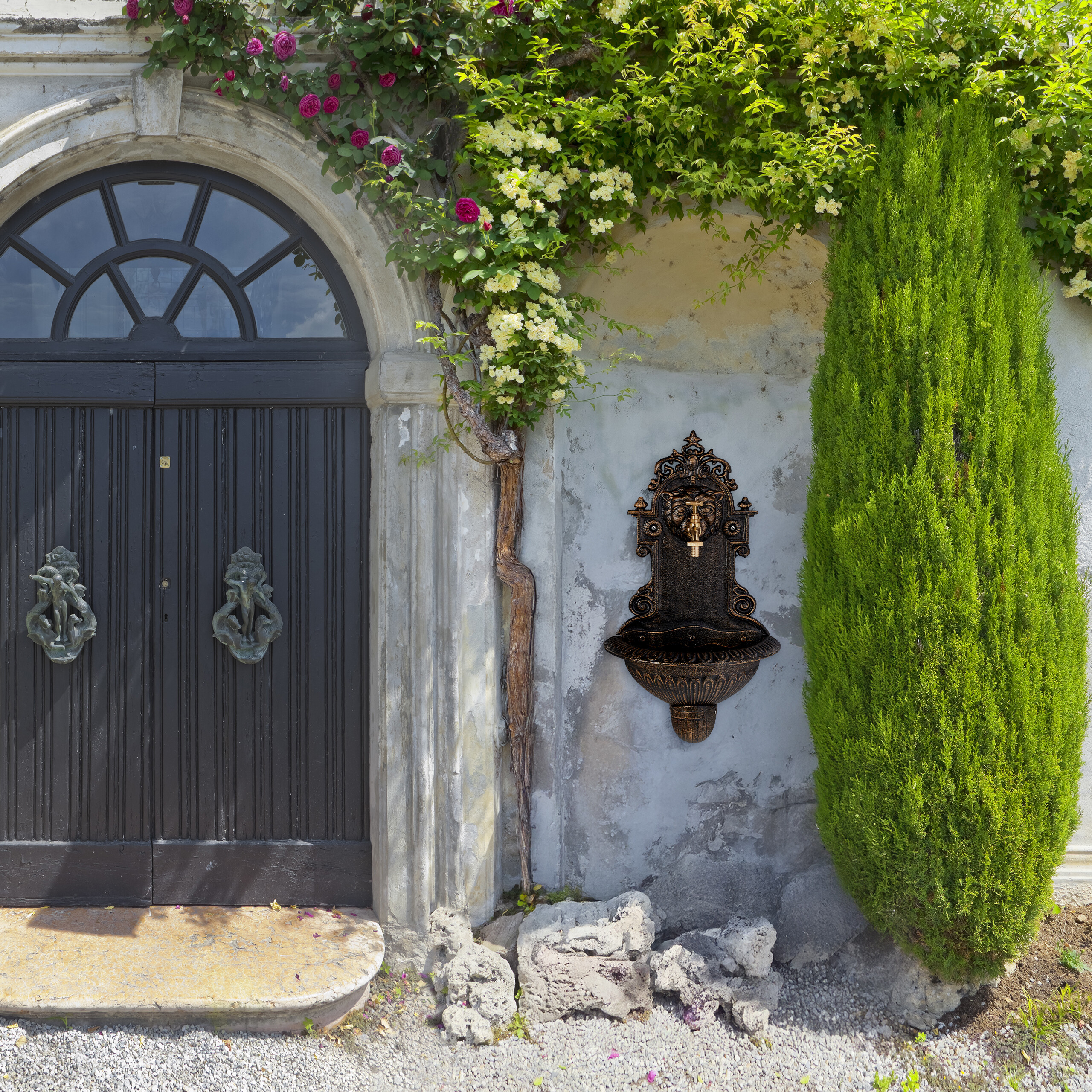 Eine schwarze Doppeltür mit Bogenfenster, ein Wandbrunnen, Kletterblumen und eine hohe grüne Zypresse.