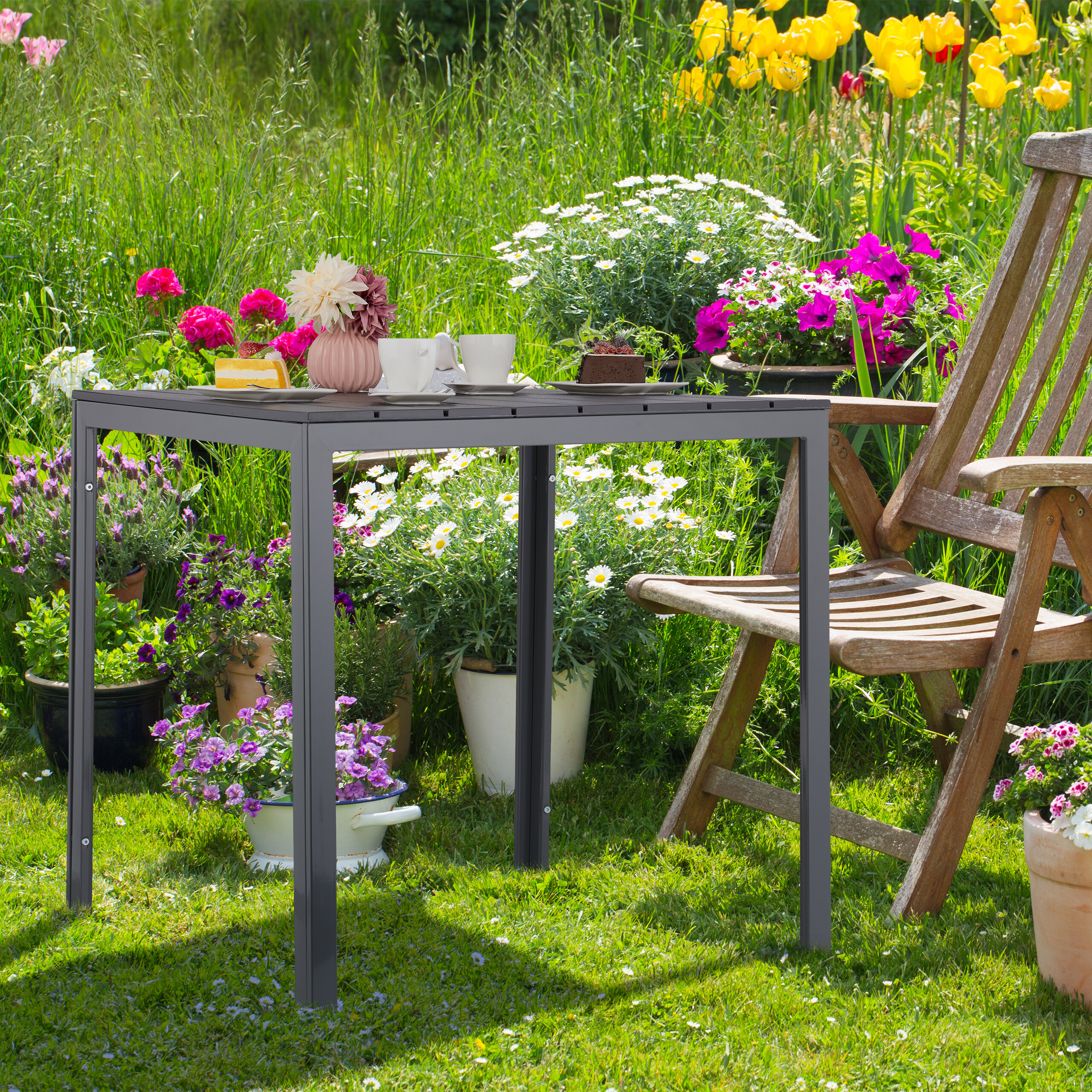 Ein grauer Gartentisch und ein Holzstuhl stehen inmitten eines blühenden Gartens.