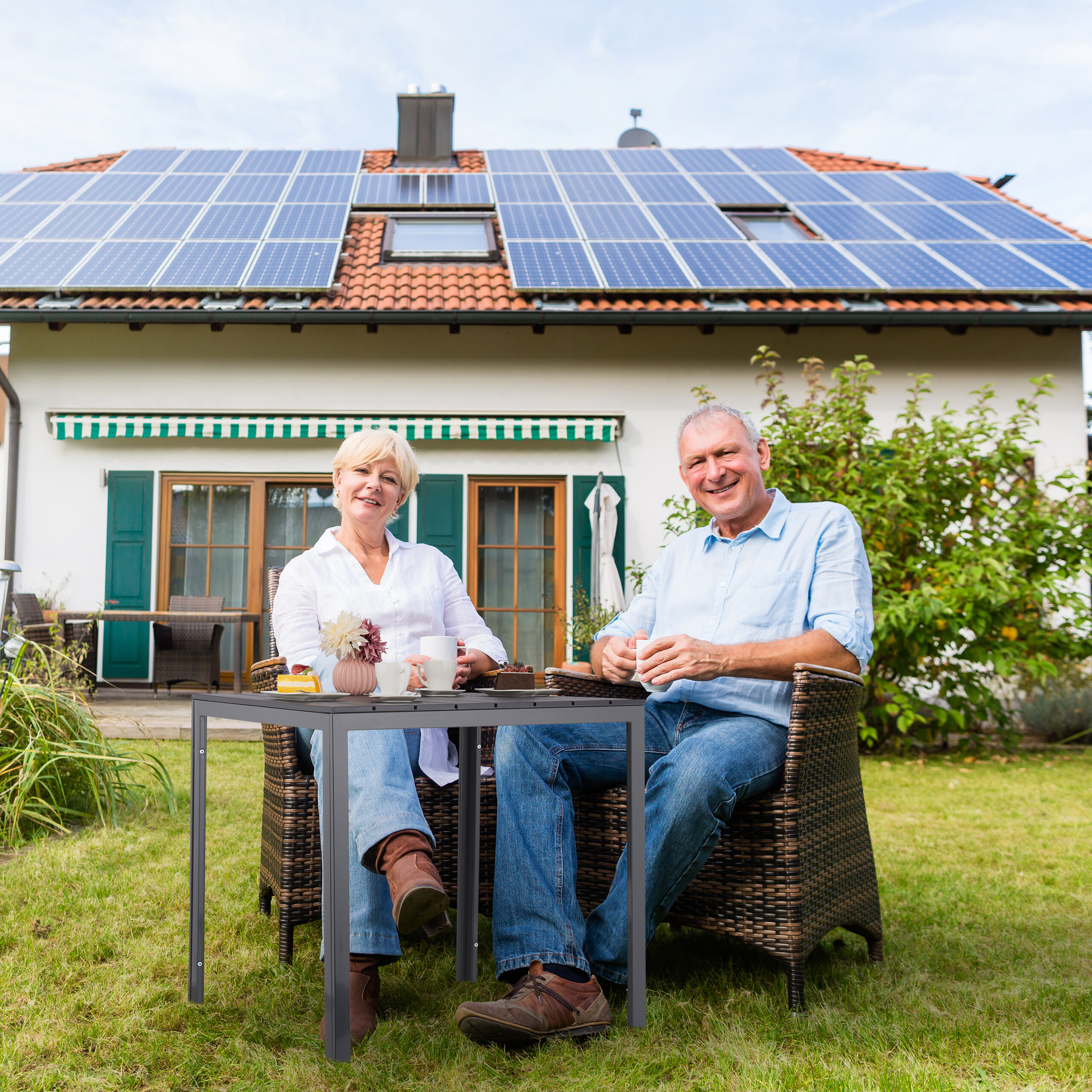 Älteres Paar sitzt entspannt im Garten vor einem Haus mit Solaranlage auf dem Dach und trinkt Kaffee.