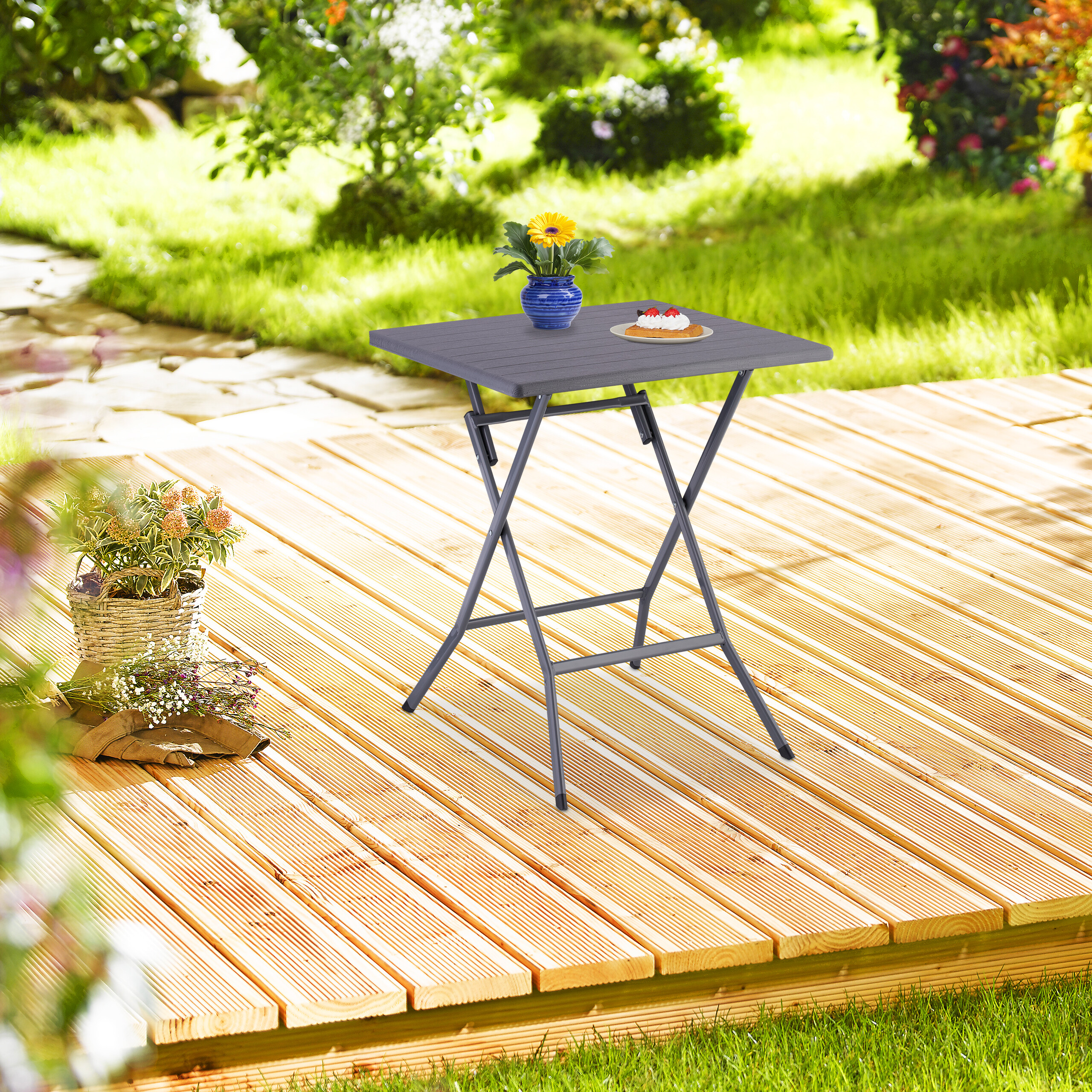 Ein grauer Klapptisch steht auf einer Holzterrasse im Garten, darauf eine Vase und ein Teller mit Kuchen.