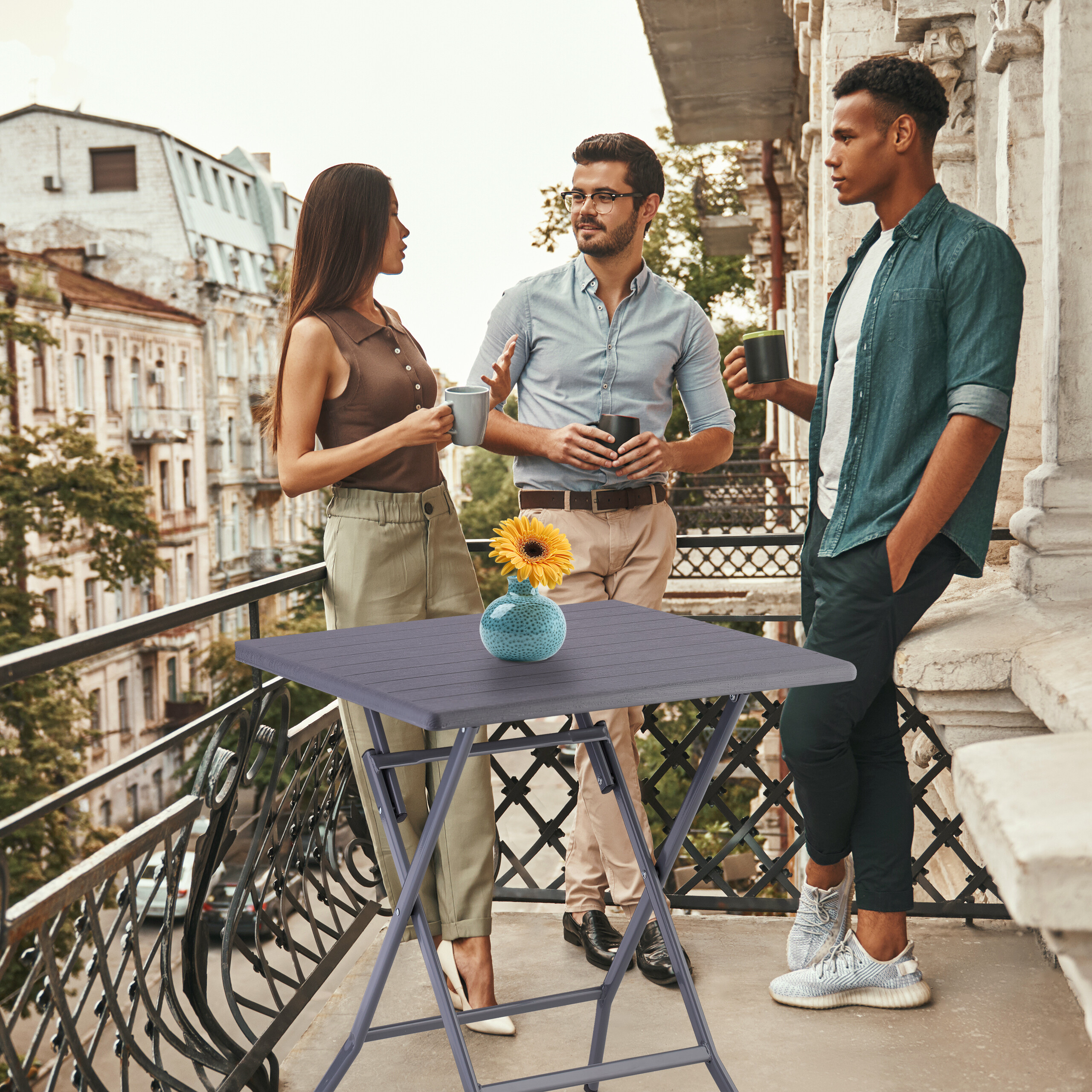 Drei Freunde stehen auf einem Balkon, halten Tassen und unterhalten sich neben einem kleinen Tisch mit einer Blume.