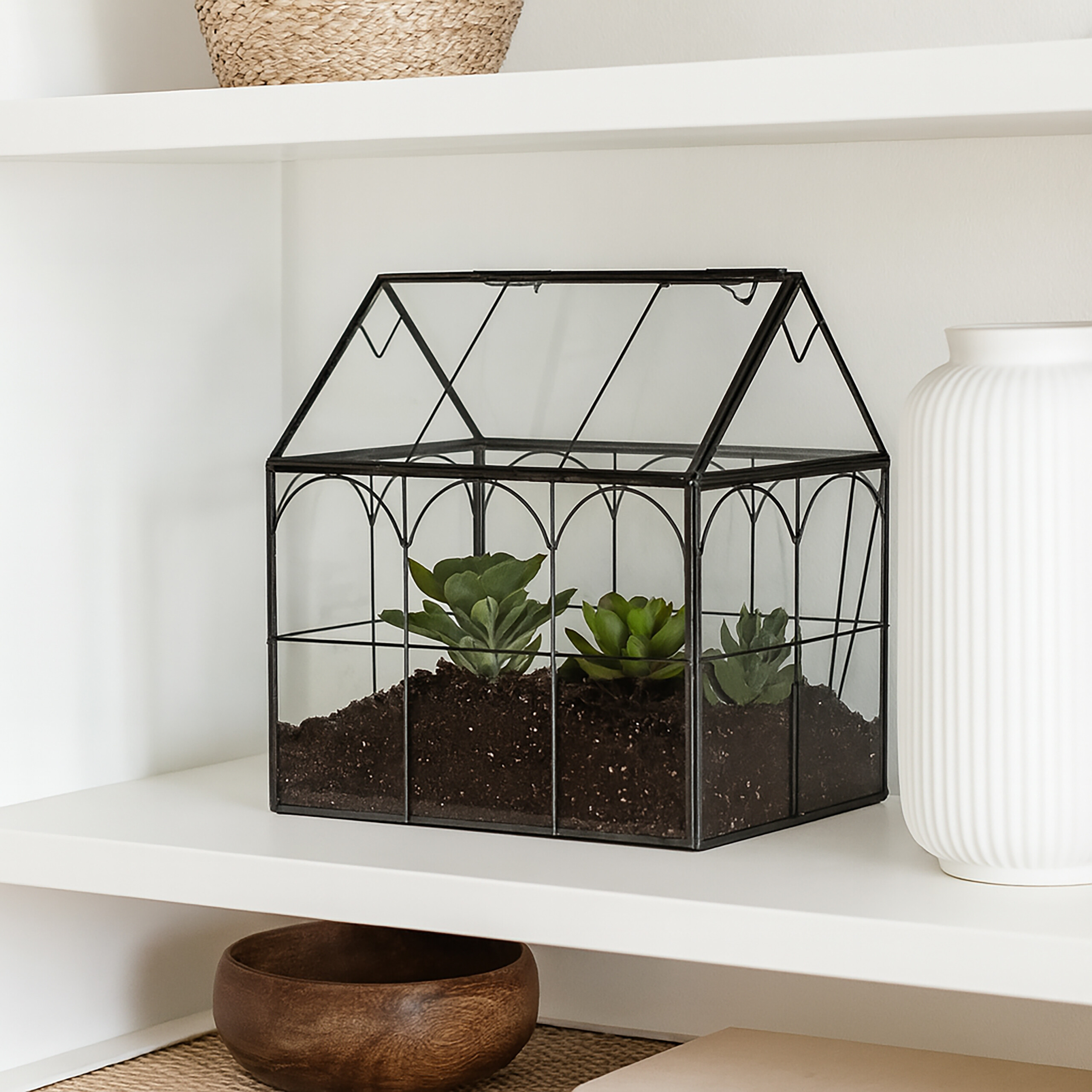 Ein schwarzes Glas-Terrarium mit Sukkulenten auf einem weißen Regal zwischen einer weißen Vase und einer Holzschale.
