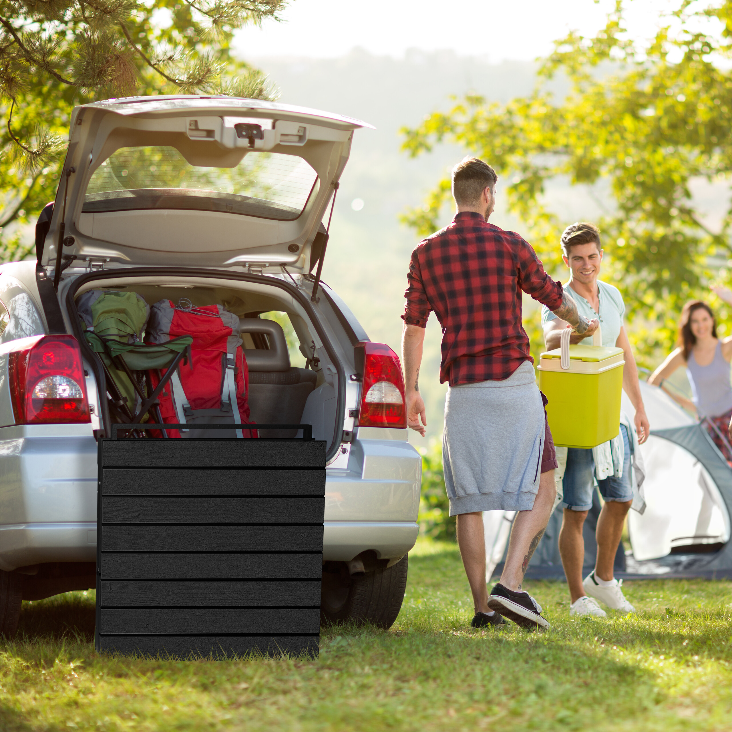 Zwei Männer packen Ausrüstung aus dem Kofferraum eines Autos auf einem Campingplatz, im Hintergrund ein Zelt und eine Frau.