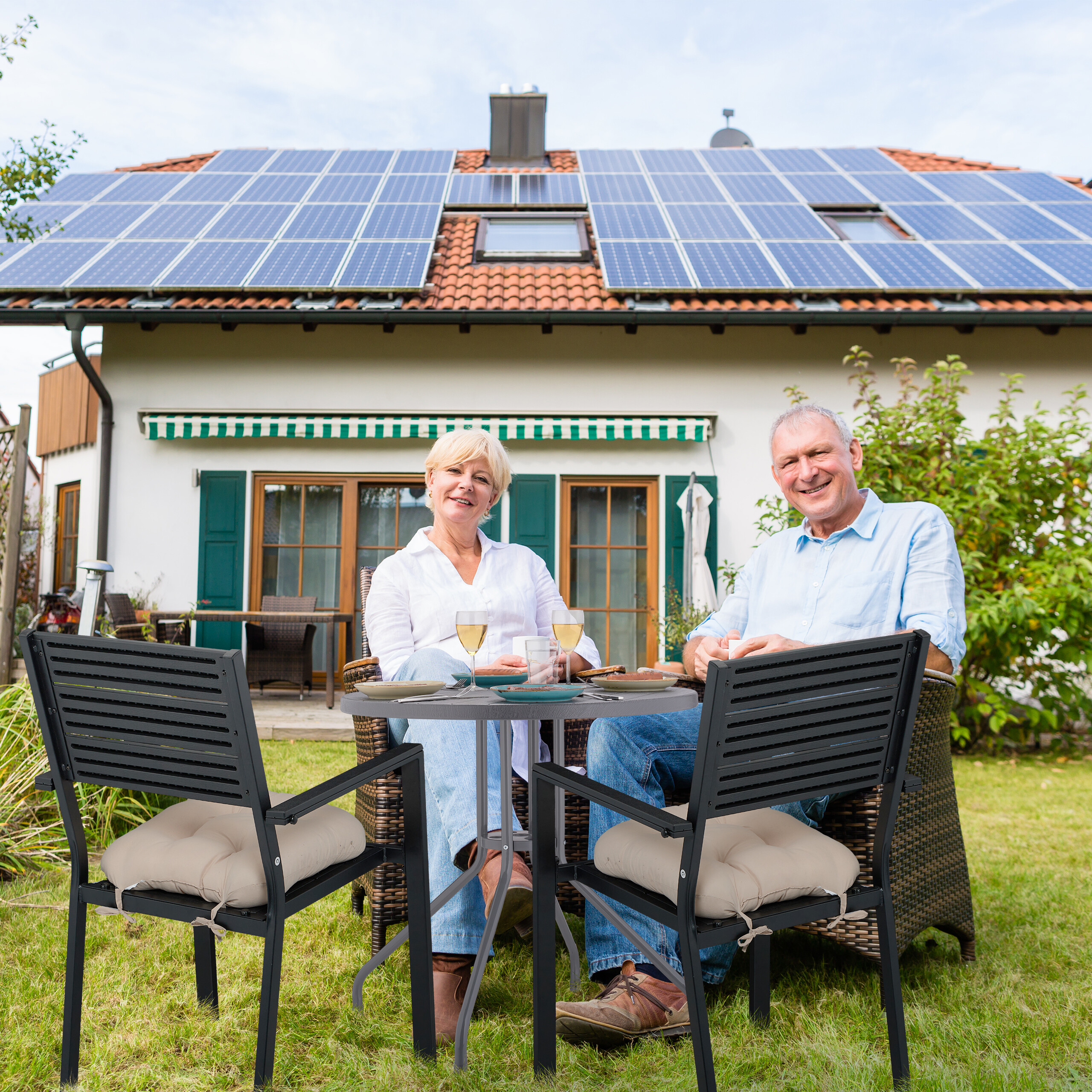 Ein älteres Paar sitzt an einem Gartentisch mit Getränken vor einem Haus mit Solaranlagen.