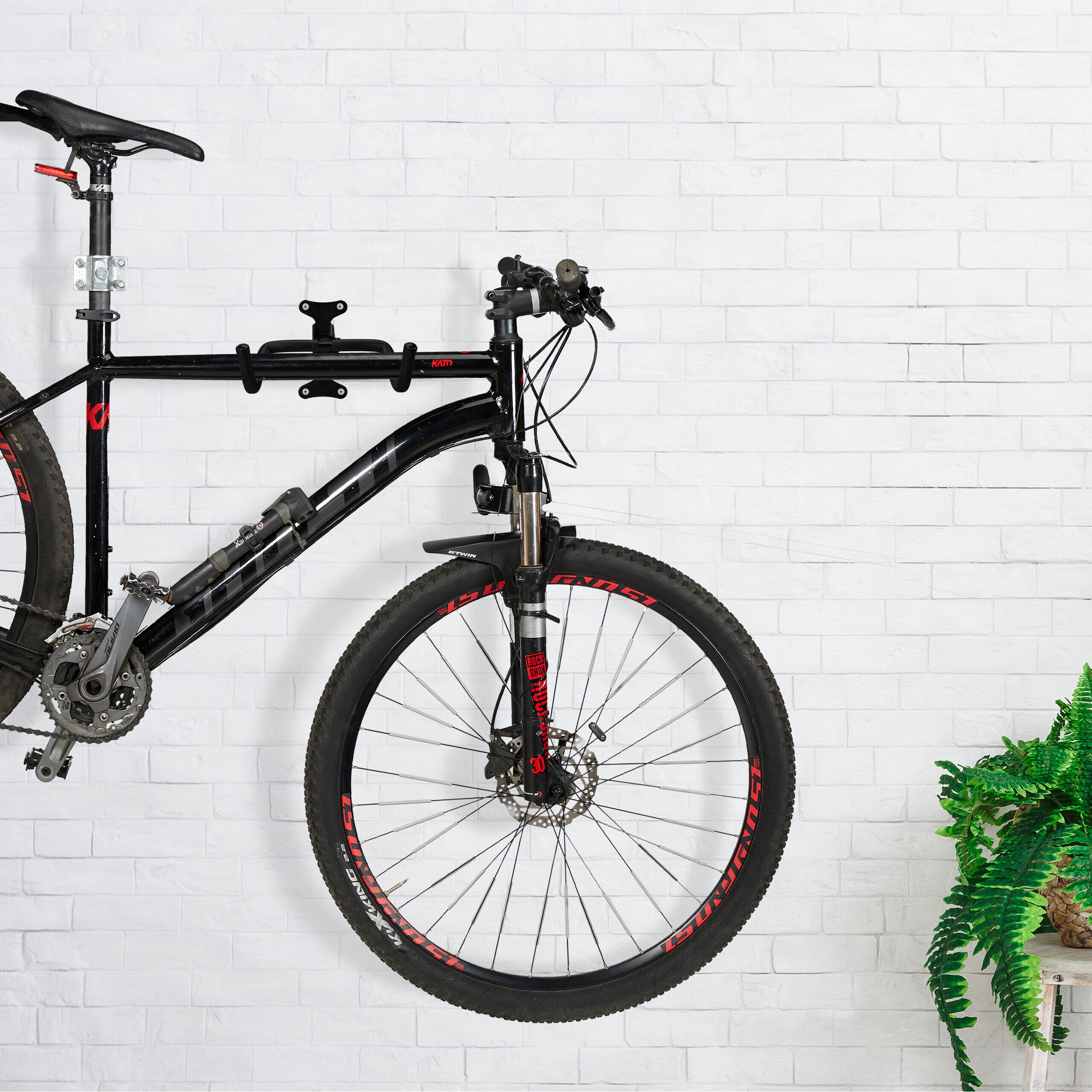 Ein schwarzes Mountainbike hängt an einer Halterung vor einer weißen Backsteinwand, rechts eine grüne Pflanze.