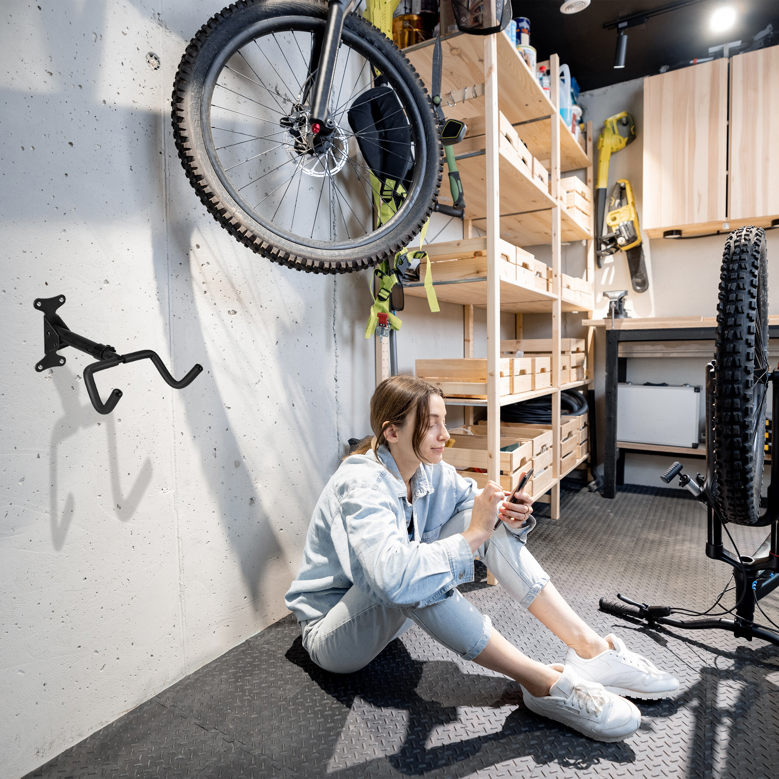 Eine Frau sitzt in einer Garage auf dem Boden und nutzt ihr Smartphone vor Werkstattregalen und einem Fahrrad.