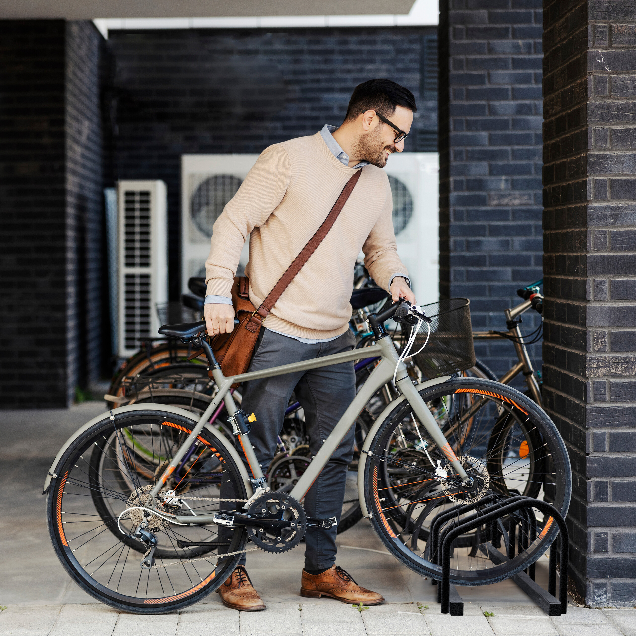 Ein Mann im beige Pullover stellt ein graues Fahrrad in einen Ständer vor einer Ziegelwand.