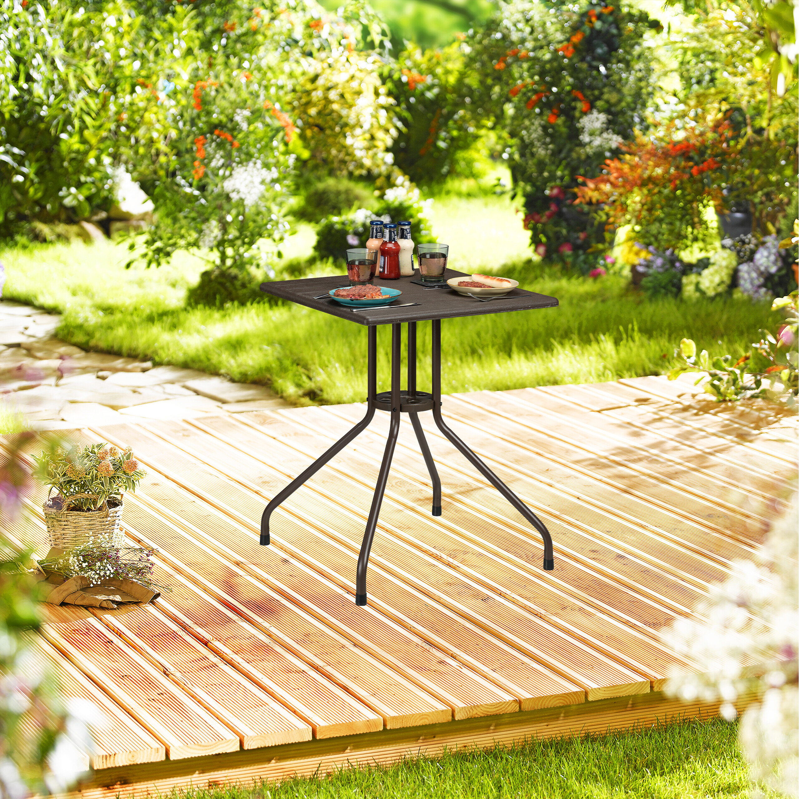 Ein kleiner dunkler Metalltisch mit Tellern, Gläsern und Gewürzflaschen steht auf einer Holzterrasse im Garten.