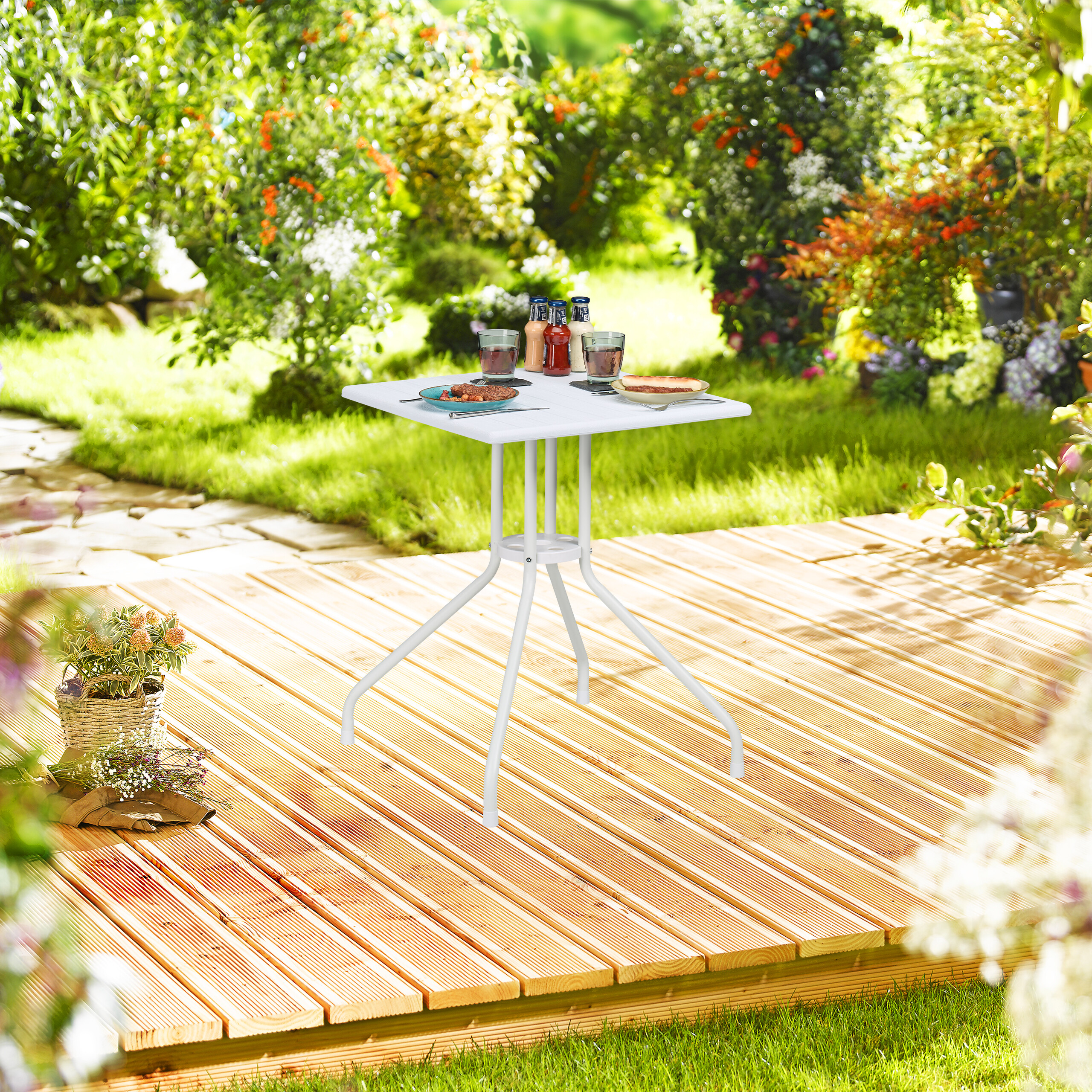 Ein weißer Tisch auf einer Holzterrasse im sonnigen Garten mit Gläsern und Essen.