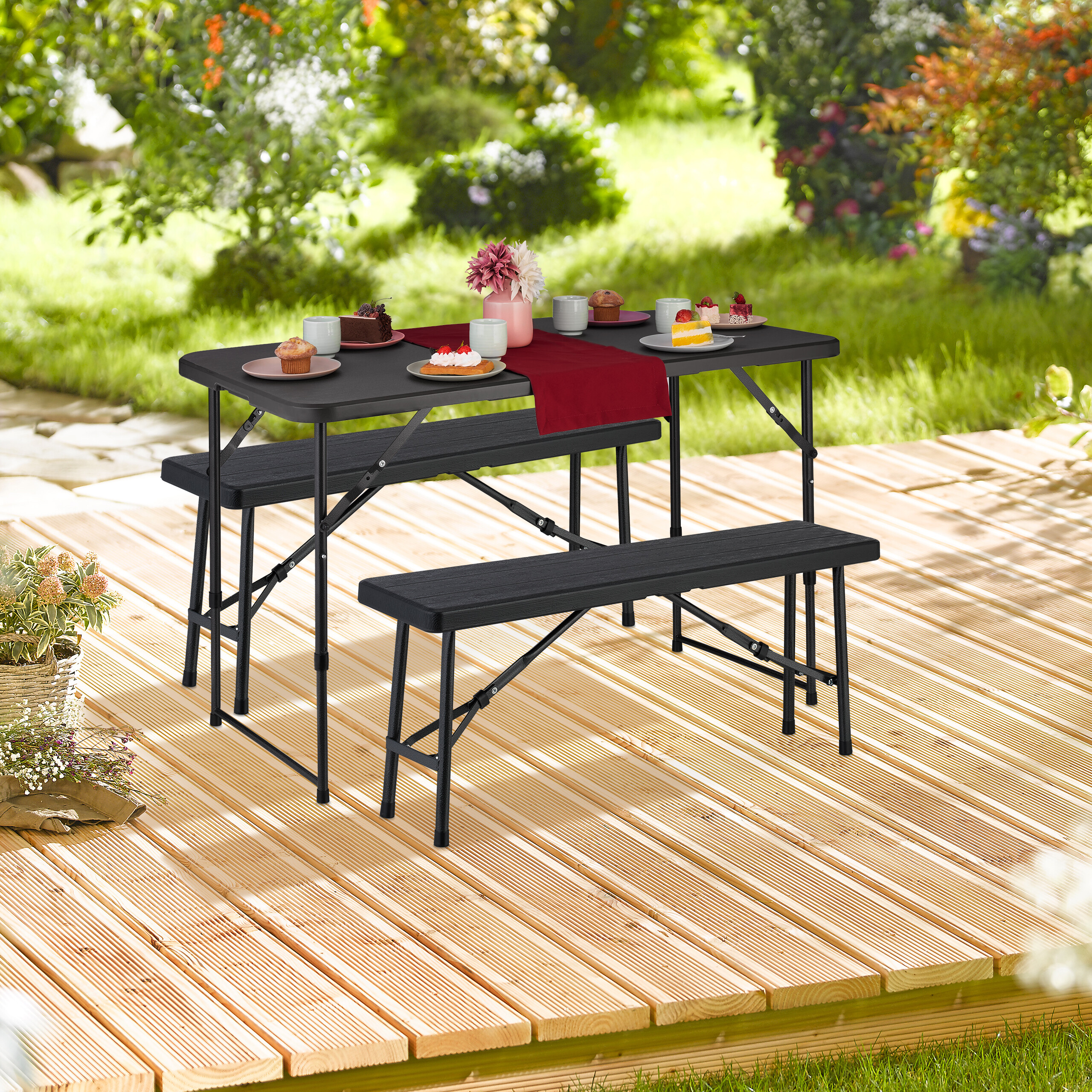 Ein schwarzer Klapptisch mit zwei Bänken steht auf einer Holzterrasse im Garten, gedeckt mit Kuchen und Blumen.