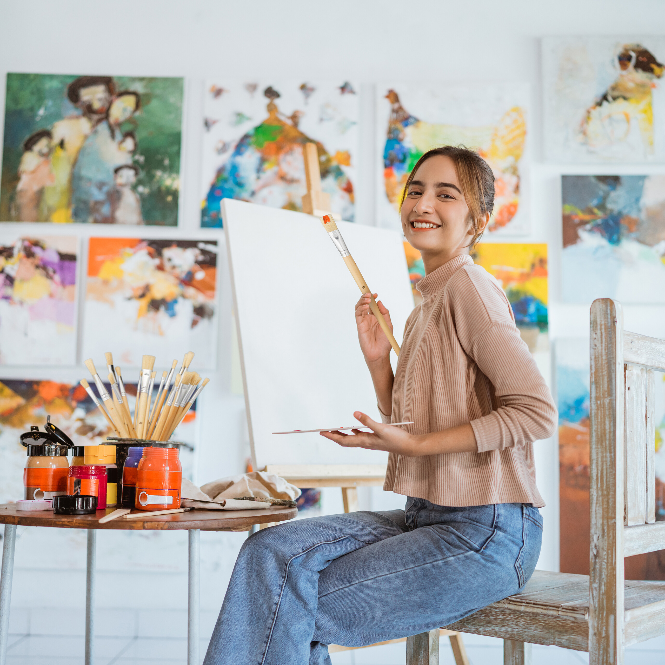 Lächelnende Künstlerin sitzt in Atelier mit Gemälden im Hintergrund und hält Pinsel und Palette.