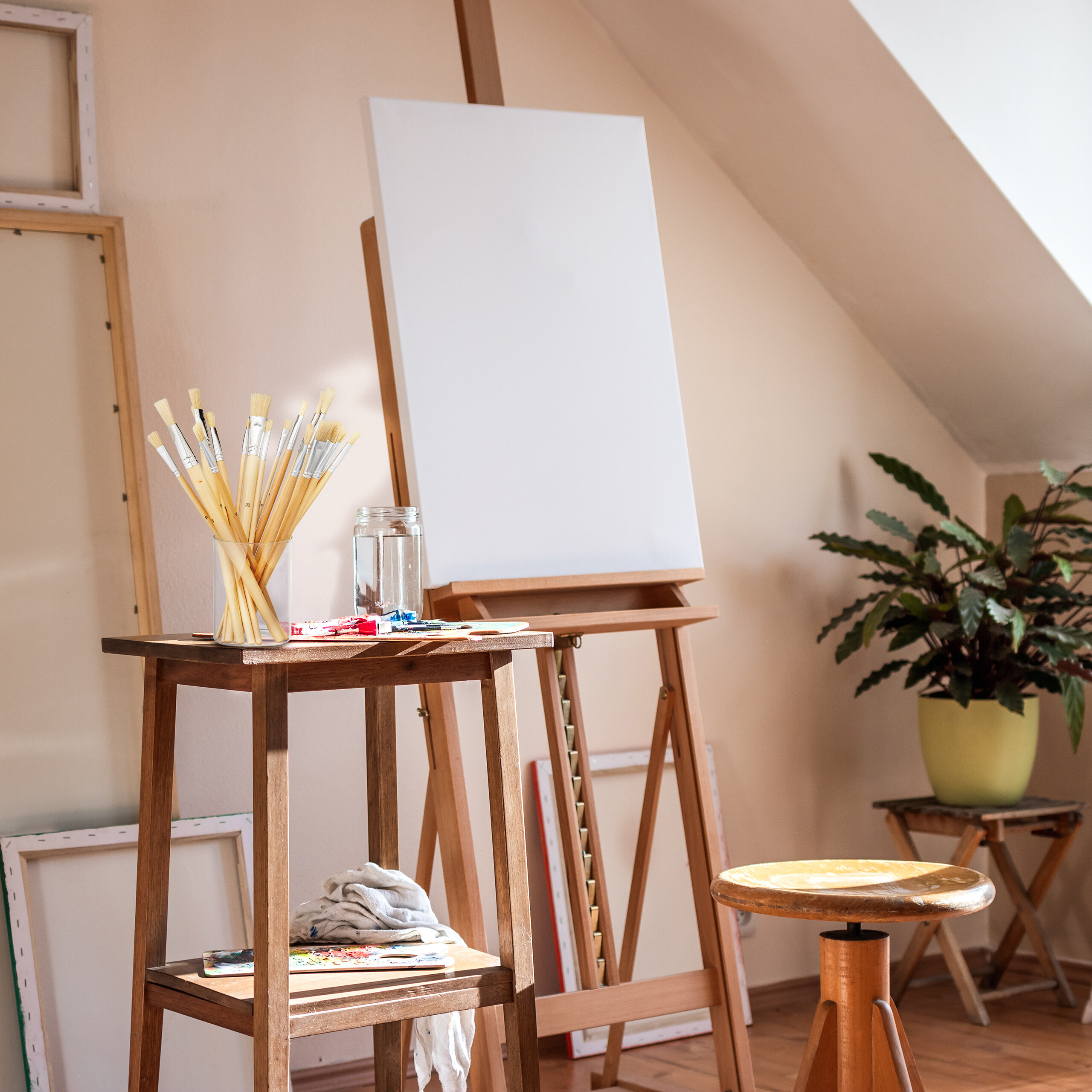 Ein Künstleratelier mit Staffelei, leerer Leinwand, Pinsel in einem Glas auf Holztisch und Topfpflanze.