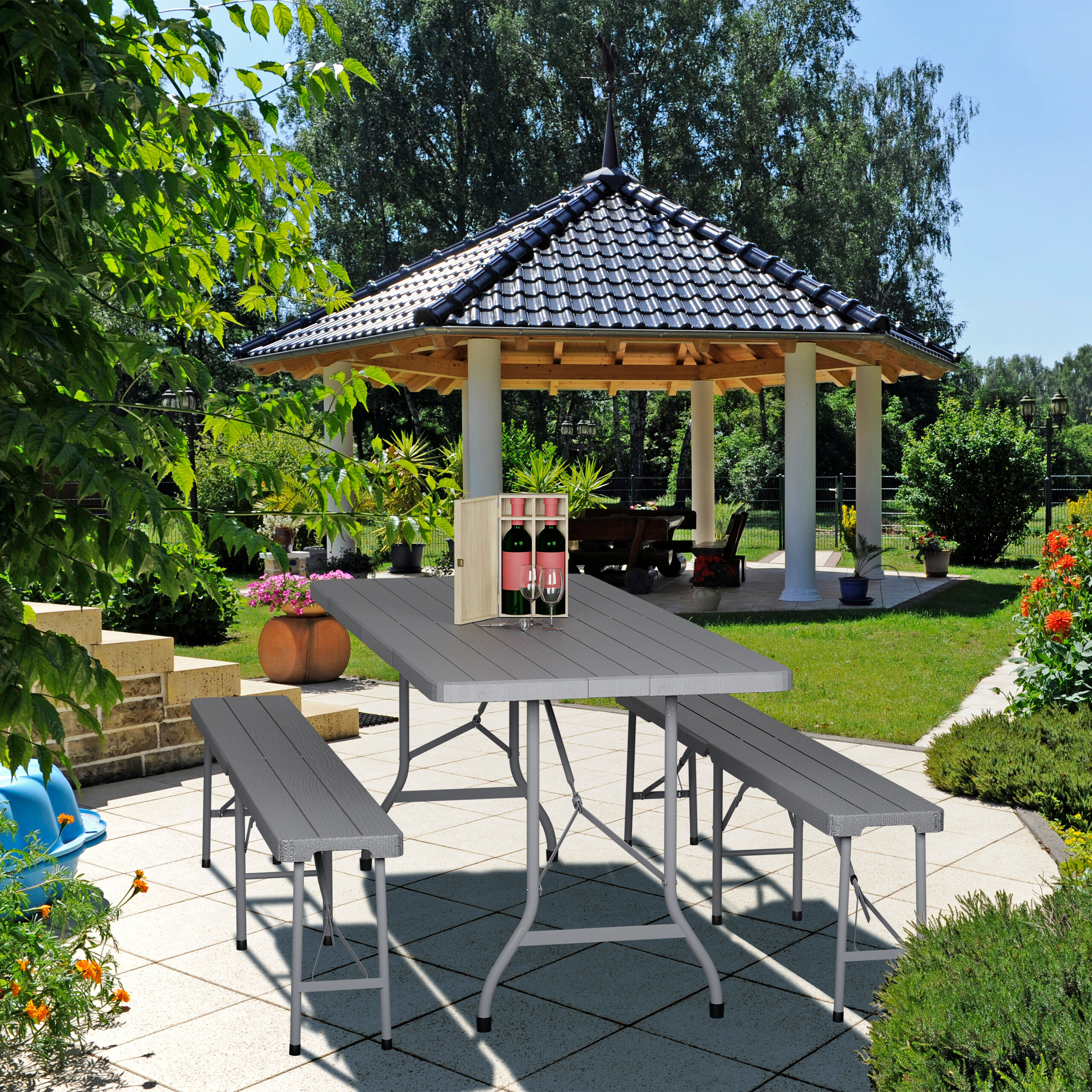 Ein grauer Klapptisch und zwei Bänke stehen auf einer sonnigen Terrasse, darauf eine Weinkiste und Gläser.