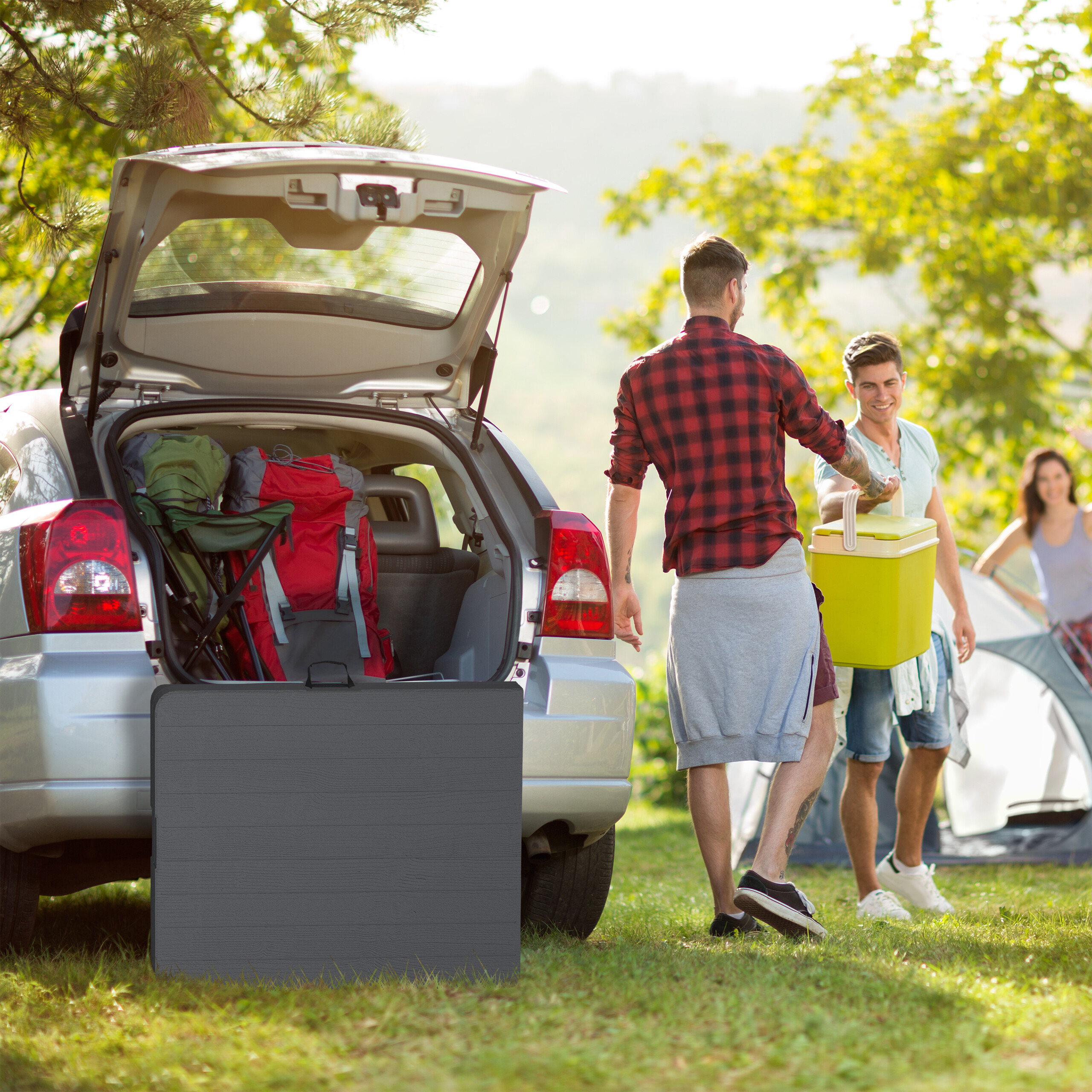 Freunde beim Campen an einem silbernen Auto mit offenem Kofferraum voller Ausrüstung und einer grünen Kühlbox.