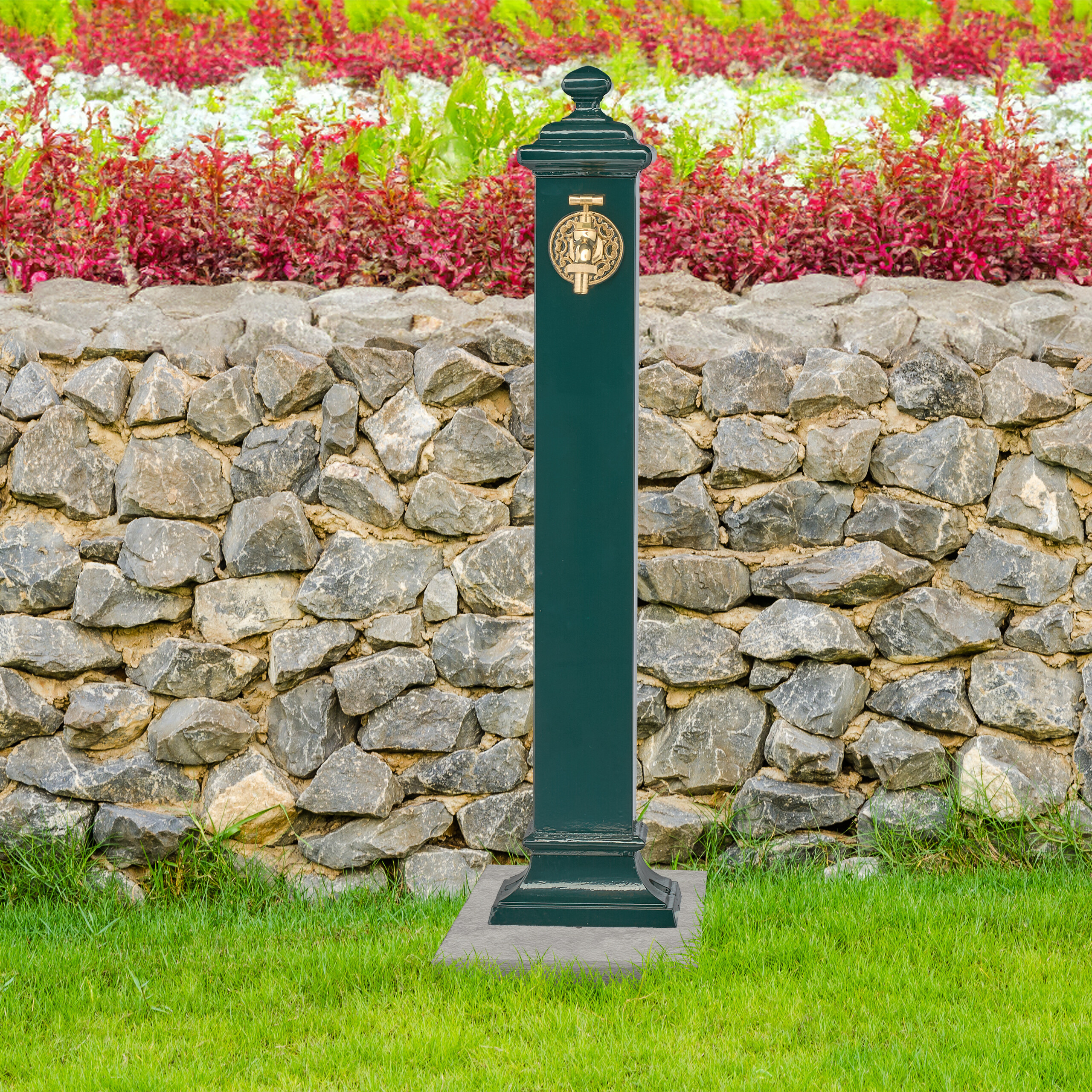 Grüner Brunnen vor einer Steinmauer und roten Blumen im Garten.