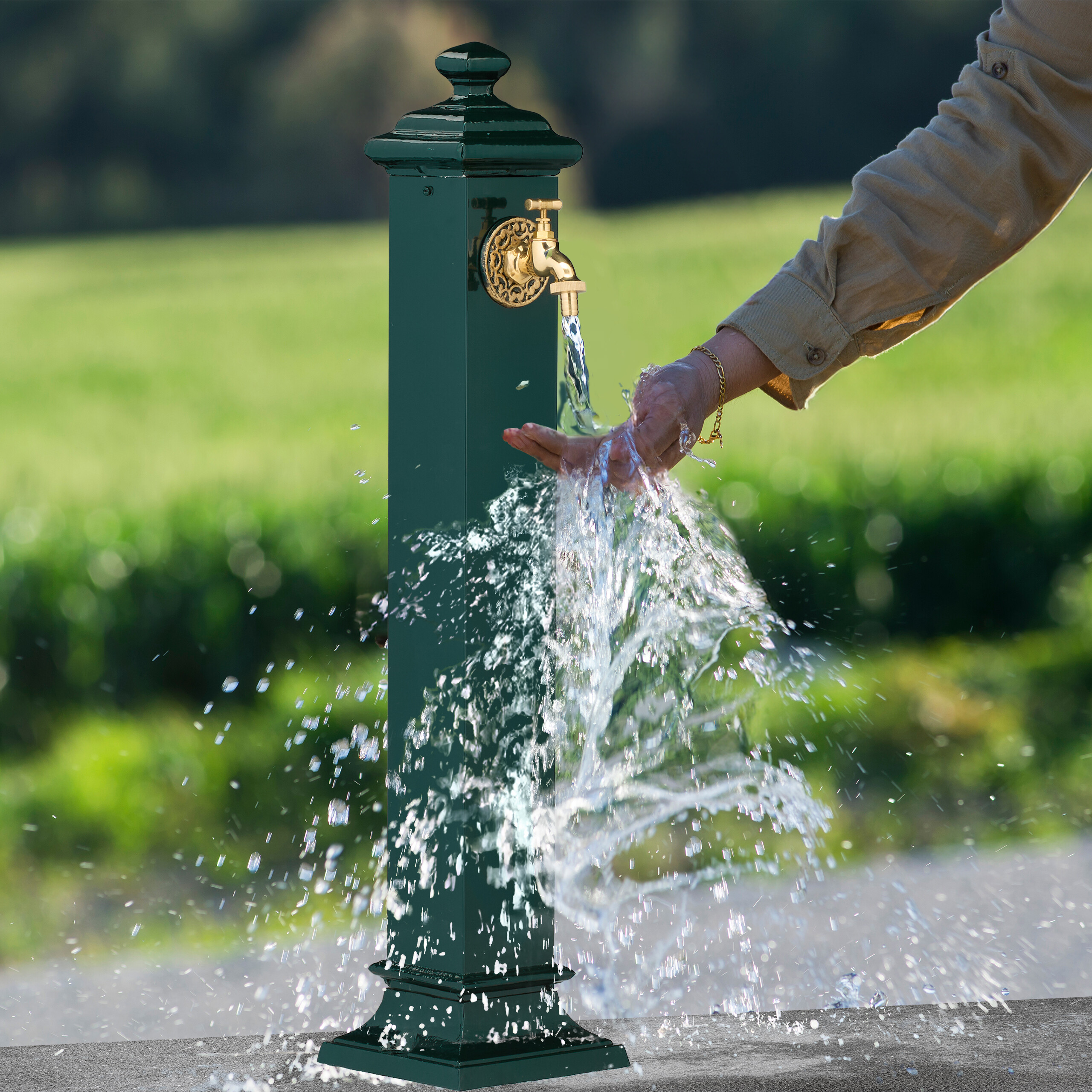 Eine Person wäscht Hände in Wasser aus einem grünen Metallbrunnen mit goldenem Hahn vor grünem Hintergrund.
