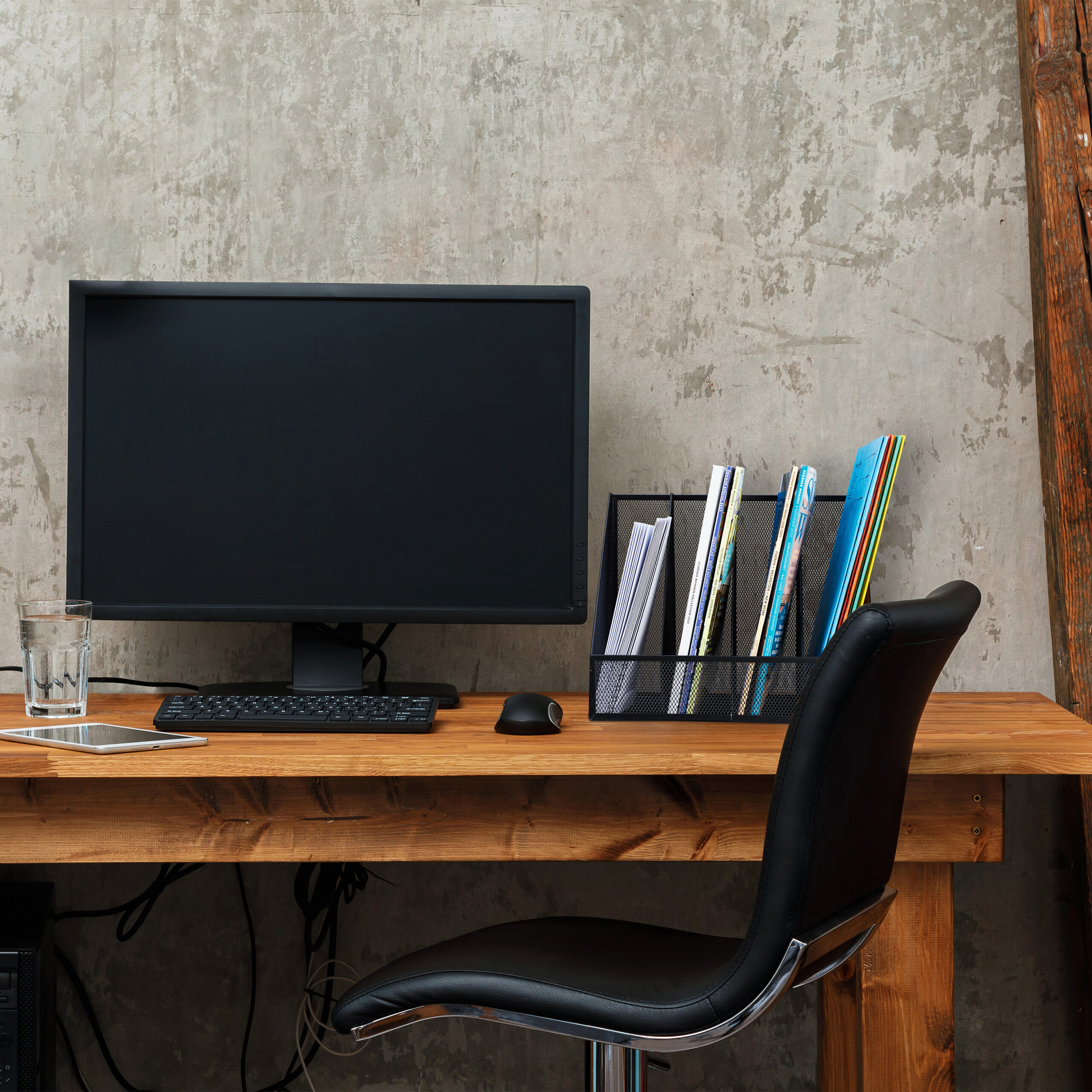 Ein Holztisch zeigt Monitor, Tastatur, Maus, Wasserglas, Tablet und Ordnerhalter vor einer Betonwand.