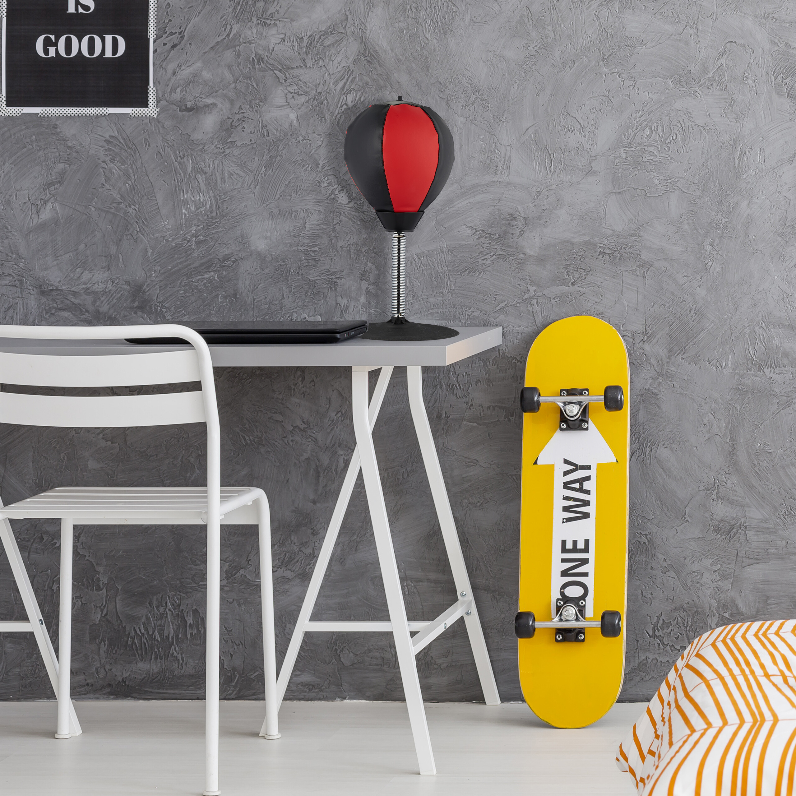 Ein weißer Stuhl, ein Schreibtisch mit Laptop, ein roter Boxball und ein gelbes Skateboard vor einer grauen Wand.